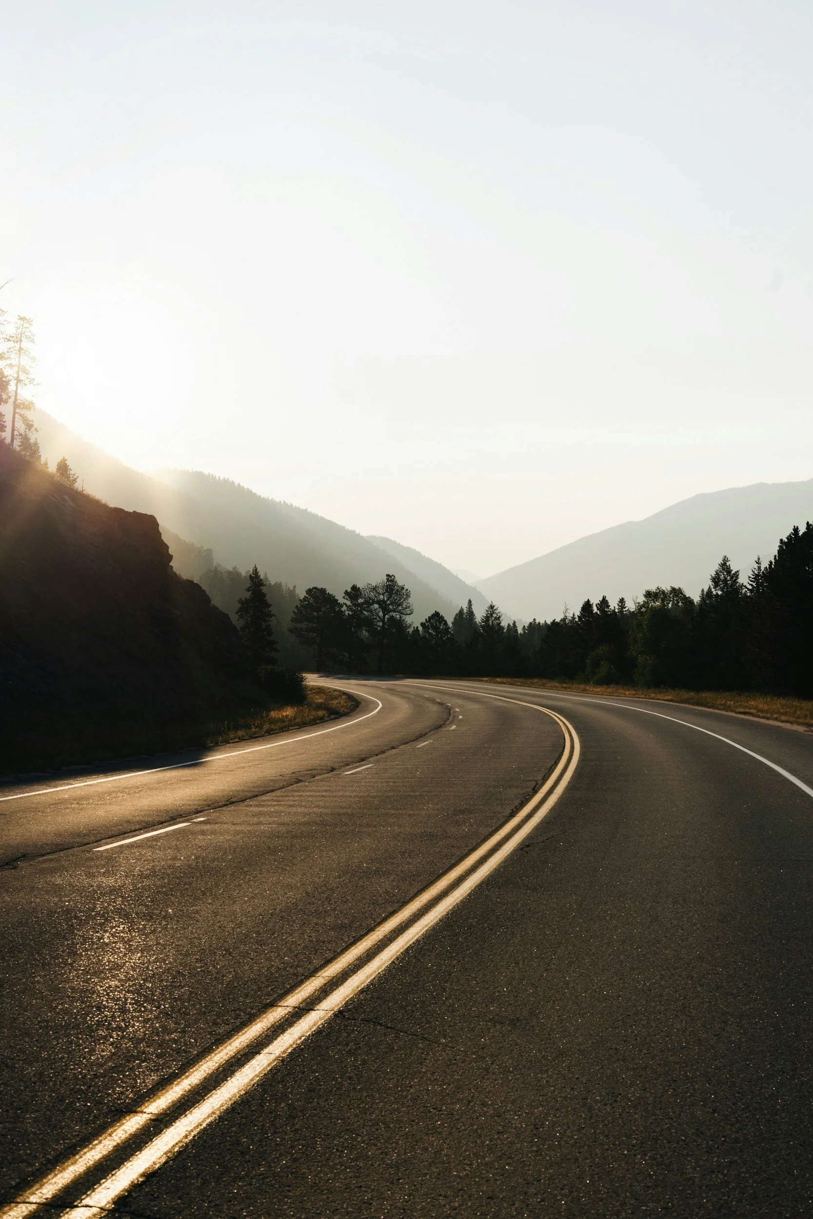 A winding mountain road at sunrise with trees and fog in the background.