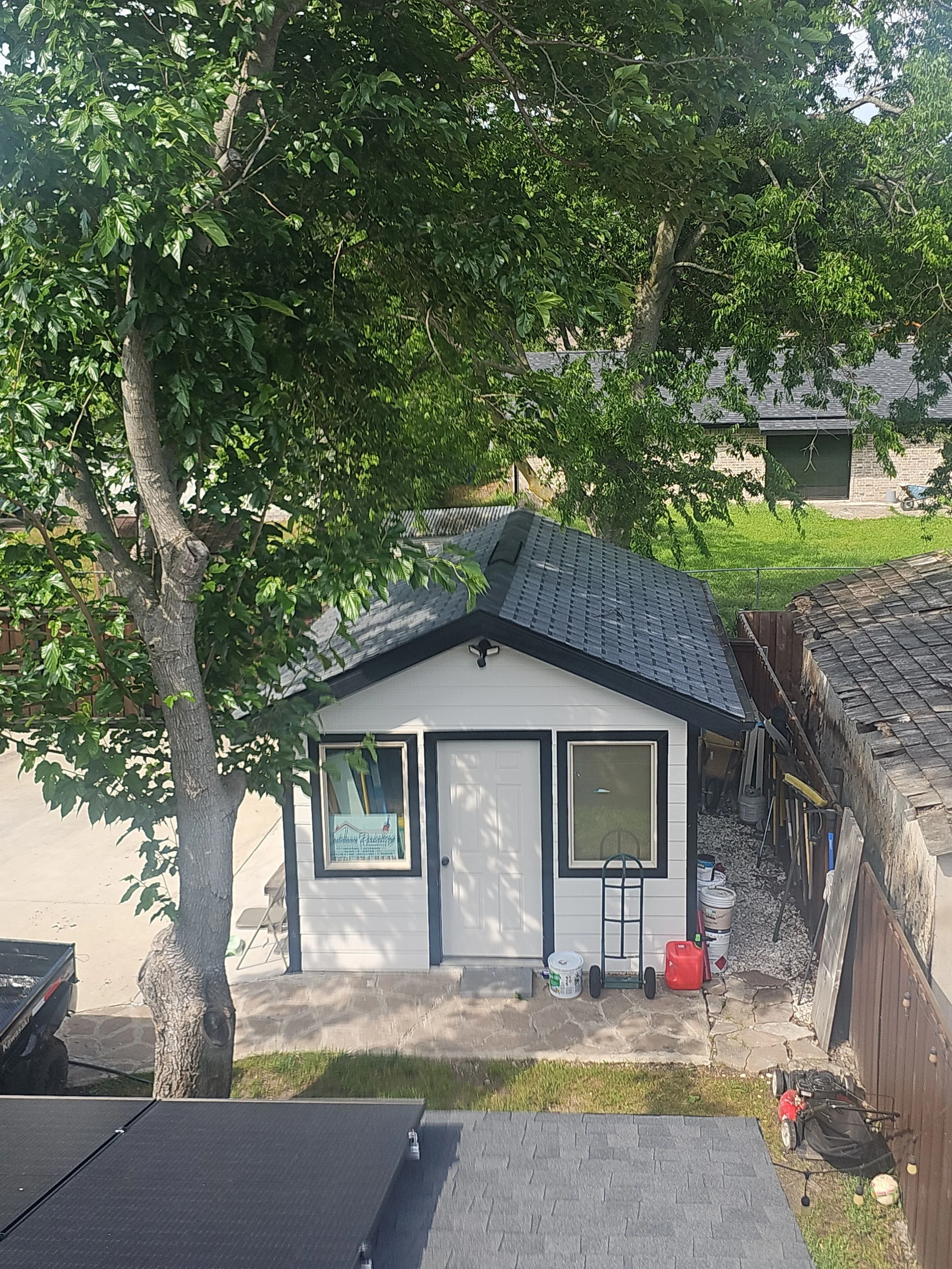A backyard with a small shed in front of a large tree. The shed has a white door and white window frames with black trim. There are buckets, a garden cart, and equipment around the shed. In the background, there is a house with a brick exterior and a
