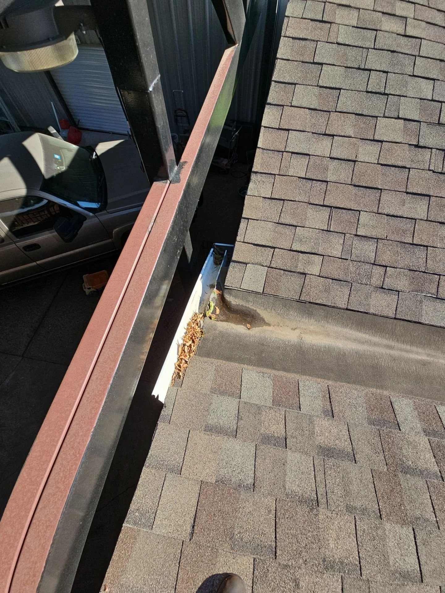 View from a rooftop showing a section of asphalt shingles, a curb, a parked car, and a gutter with fallen leaves.