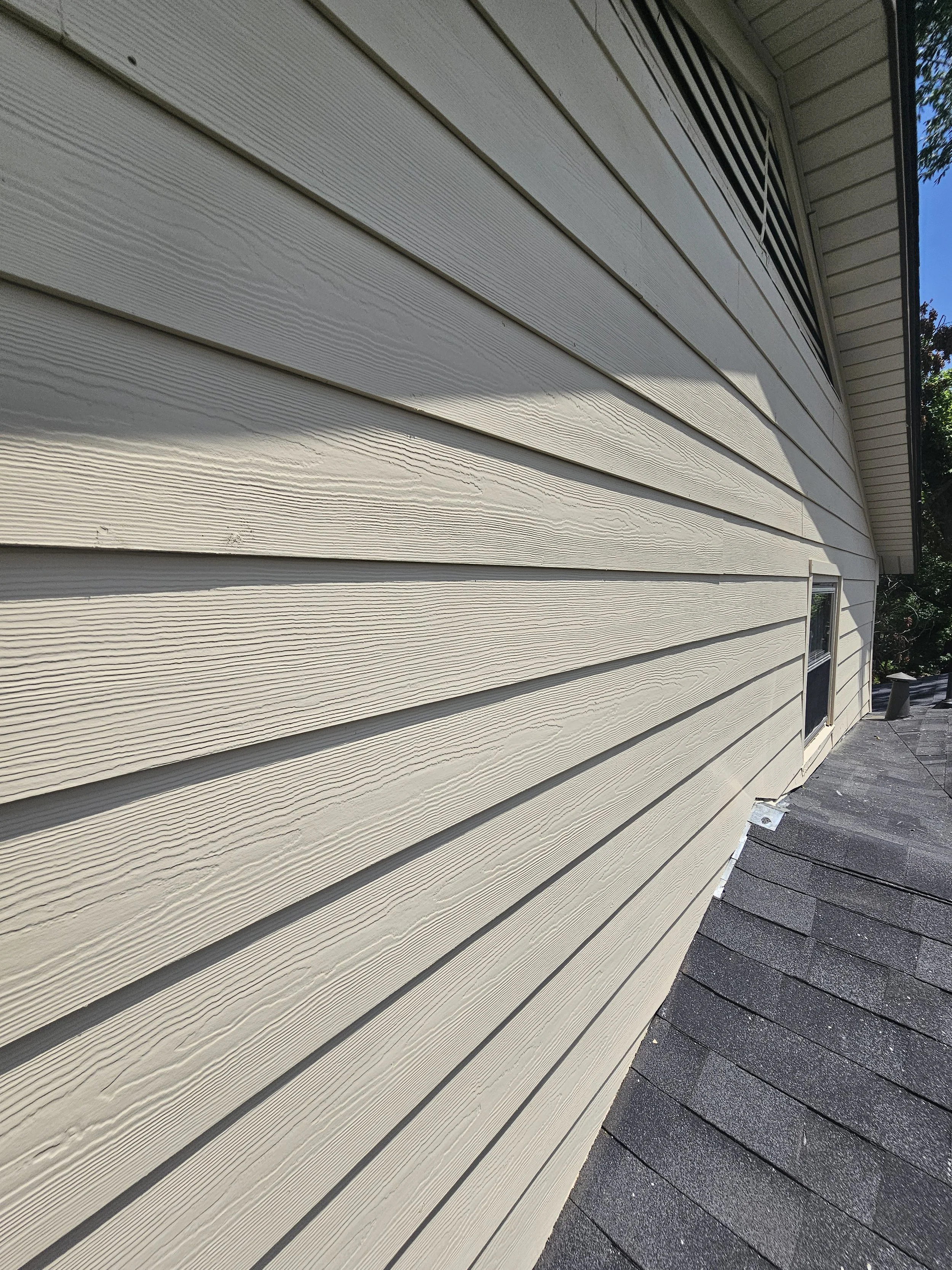 Close-up of beige horizontal siding on a house with gray asphalt shingles on the roof, sunlight casting shadows on the siding.