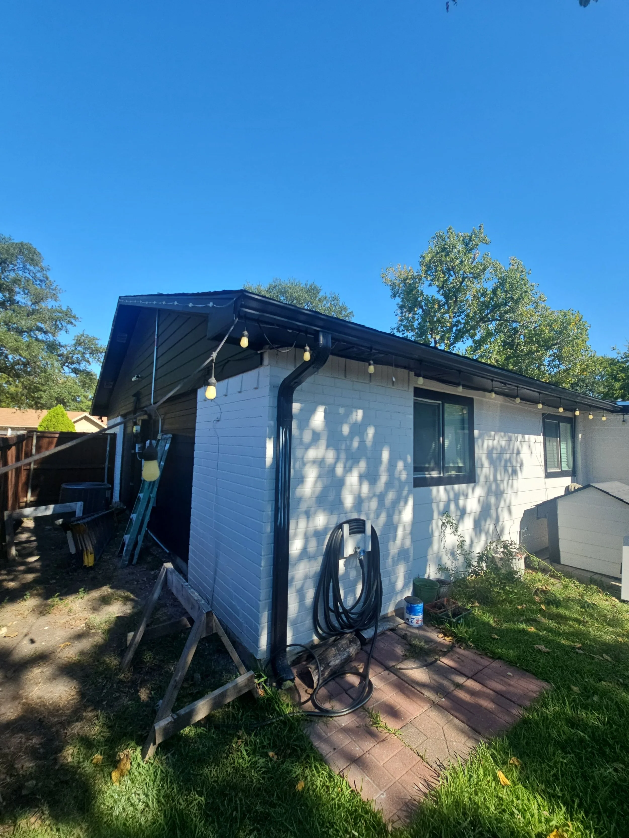 Backyard scene with part of a white house, a black gutter, outdoor string lights hanging from the roof, a garden hose on a reel, a garden area with plants and a brick pathway, and trees under a clear blue sky.