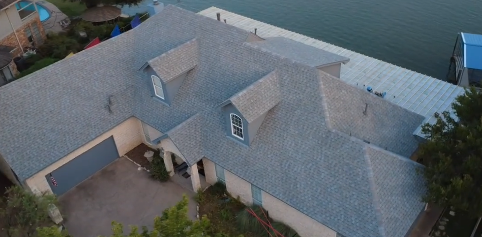 Aerial view of a house with a gray shingle roof next to a body of water, with a dock, boat cover, and surrounding trees and neighboring structures.