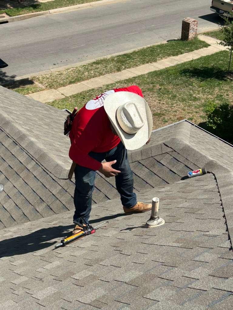 A man working on a rooftop wearing a cowboy hat, red shirt, and jeans with a tool belt, kneeling near a vent pipe, with tools and a can nearby.