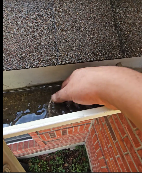 A person rinsing a sponge in a rain gutter attached to a brick house wall.