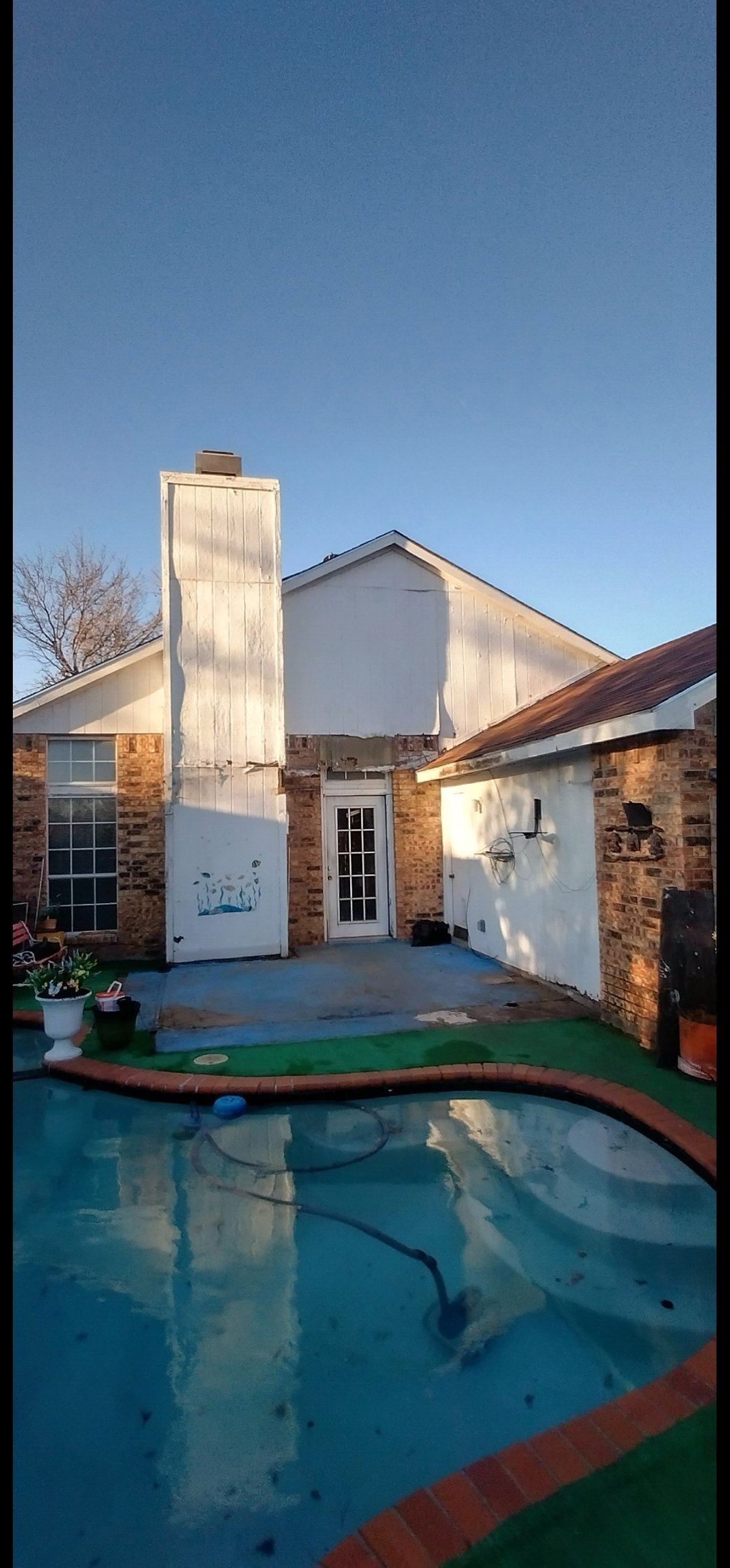 Backyard with a swimming pool, brick house, and white exterior walls under a clear blue sky.