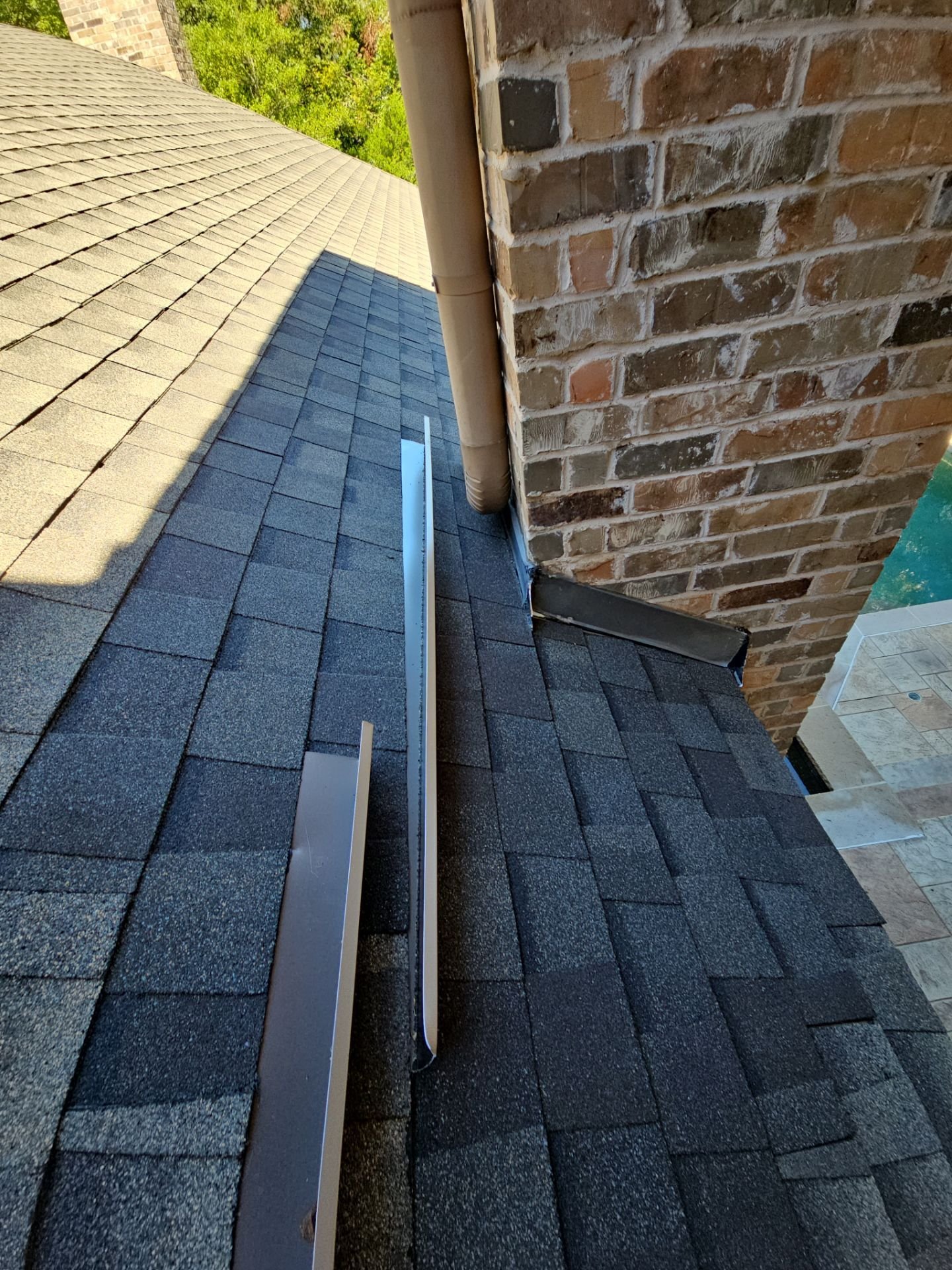 View of a house roof with asphalt shingles, a brick chimney, and a gutter pipe, with trees in the background.