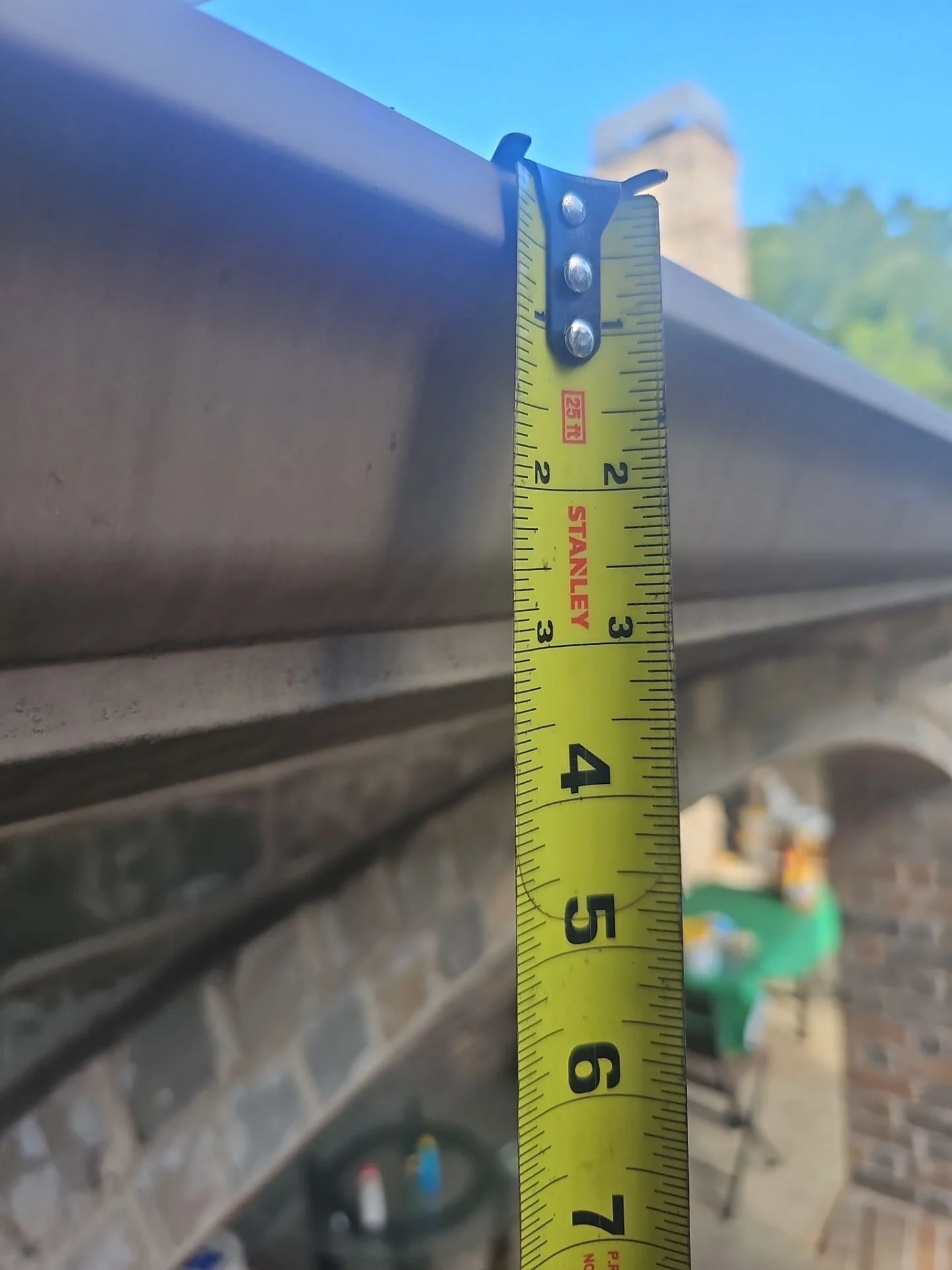 A yellow measuring tape extended vertically against a metallic surface, with a blue sky and a blurred building and trees in the background.