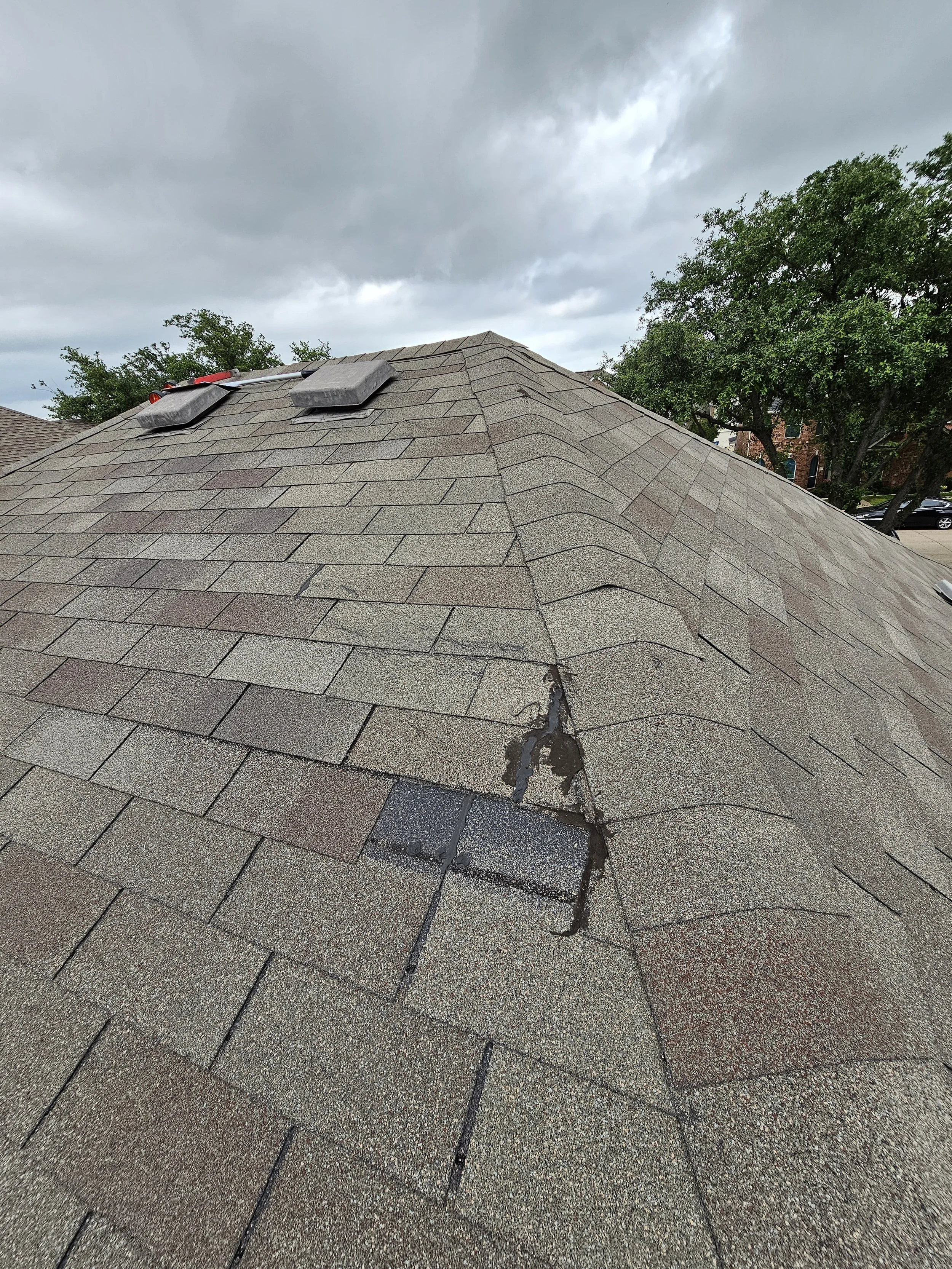 A residential roof with asphalt shingles, showing a visible crack and damage, under a cloudy sky. There are trees and neighboring houses in the background.