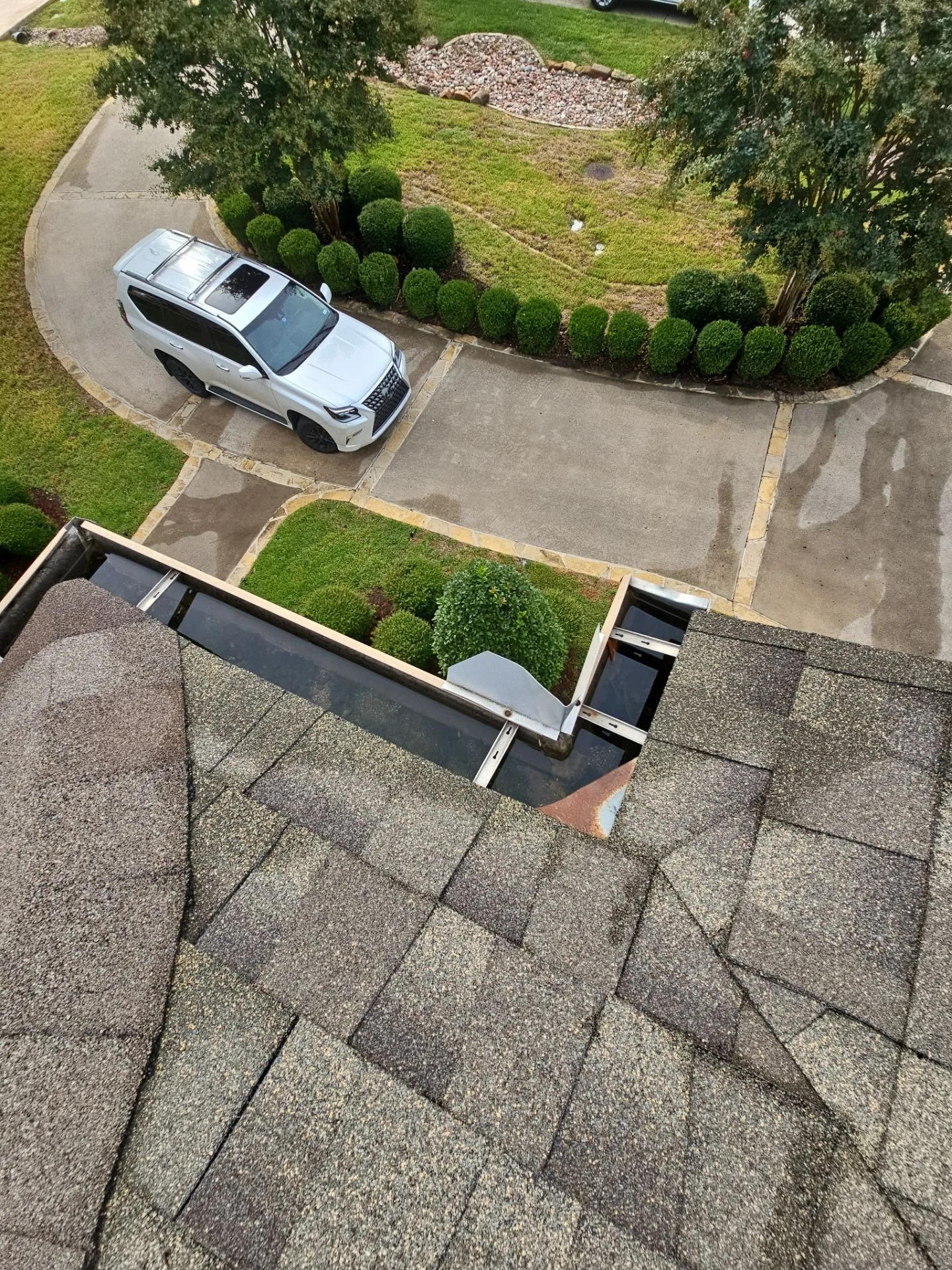 View from a balcony showing a section of roof with shingles, a gravelly parking lot with a white SUV, a landscaped garden with bushes and trees, and a sidewalk.