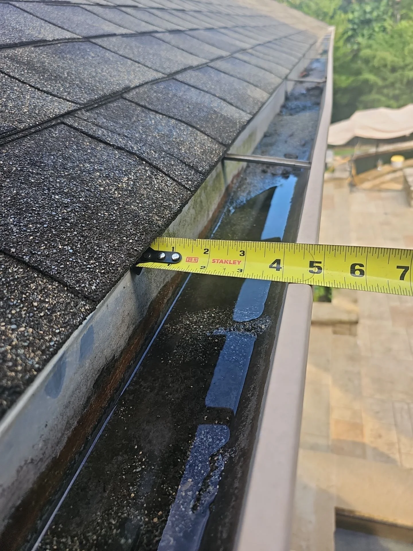 Close-up of a roof with black asphalt shingles and a measuring tape indicating about 5 inches, with a gutter underneath and a yard with trees in the background.