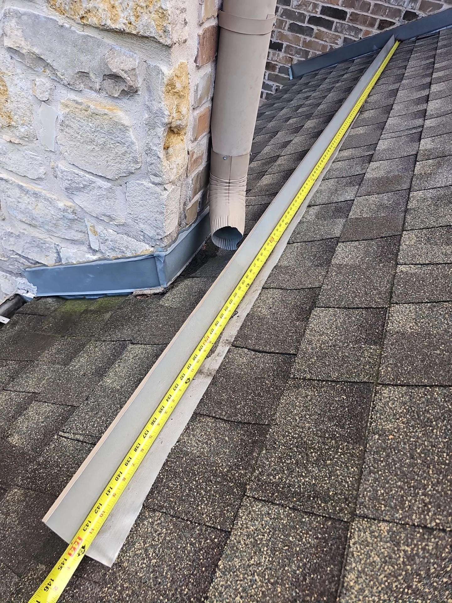 Rooftop with asphalt shingles, metal flashing, beige gutter downspout, and yellow measuring tape placed alongside a white metal vent pipe near a brick and stone exterior wall.