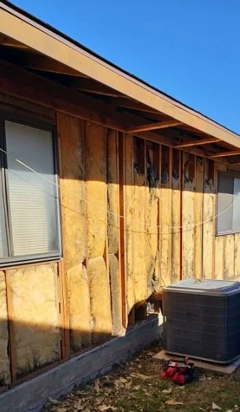 The exterior of a house with exposed wall insulation and window, with an air conditioning unit and tools on the ground.