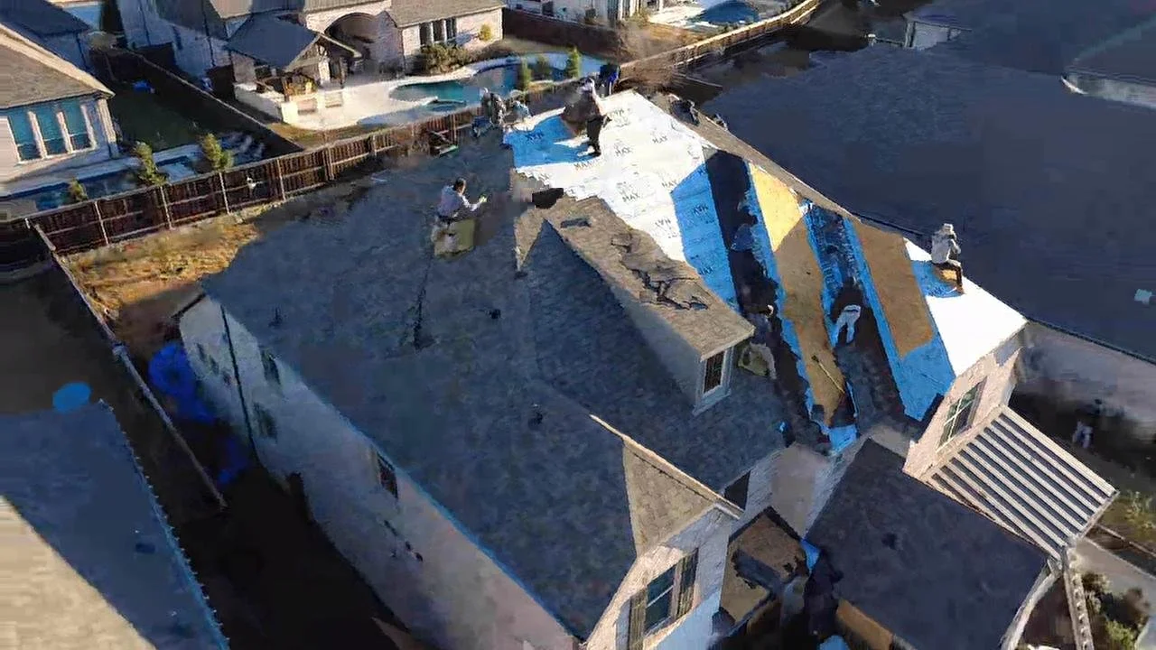 People working on a roof in a residential neighborhood, installing new roofing material. Some workers are visible near the shingles, and a backyard with a swimming pool is visible in the background.