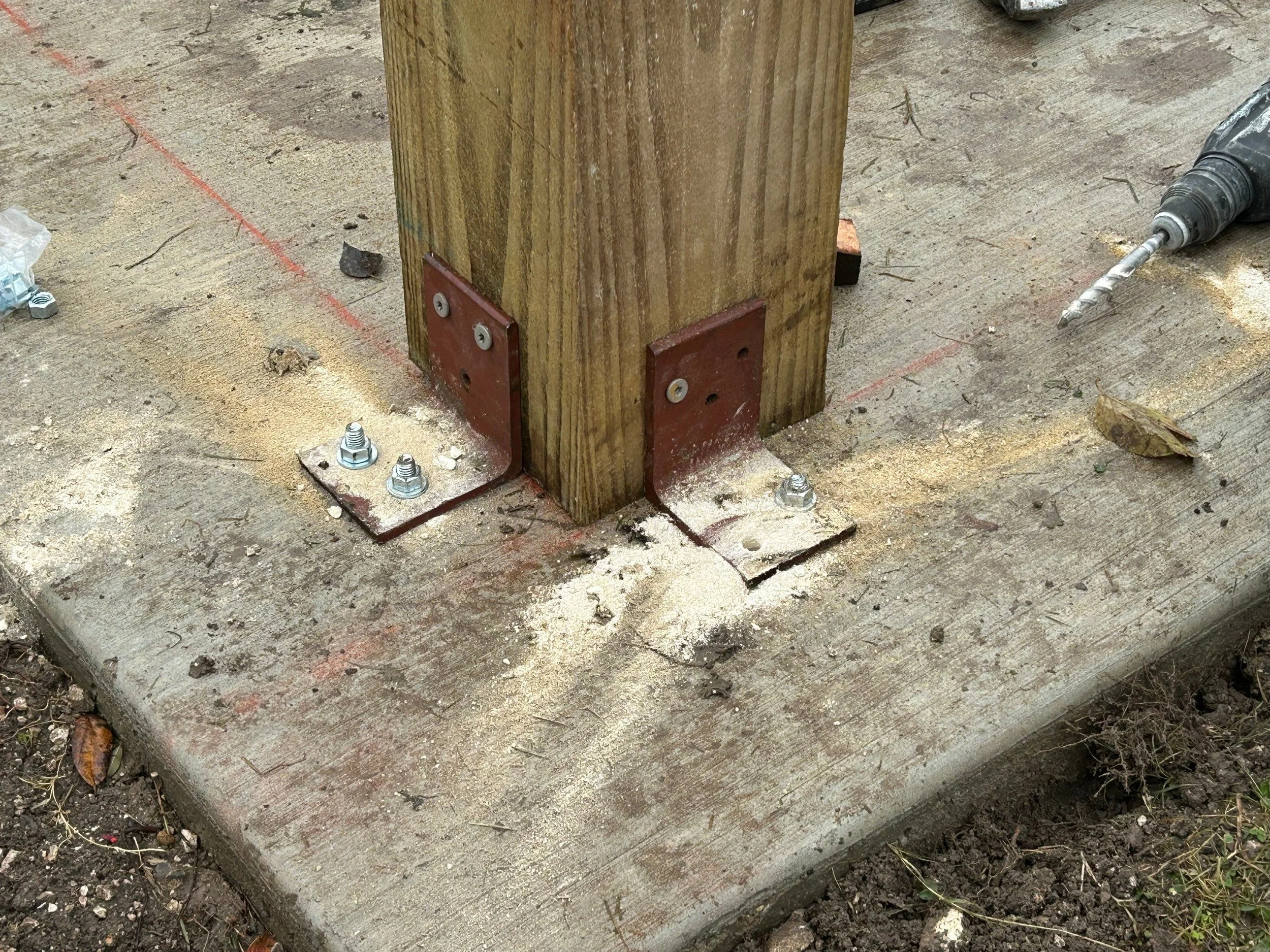 Close-up of a wooden post attached to a concrete step with metal brackets and bolts, with a cordless drill nearby, outdoor setting with dirt and leaves.