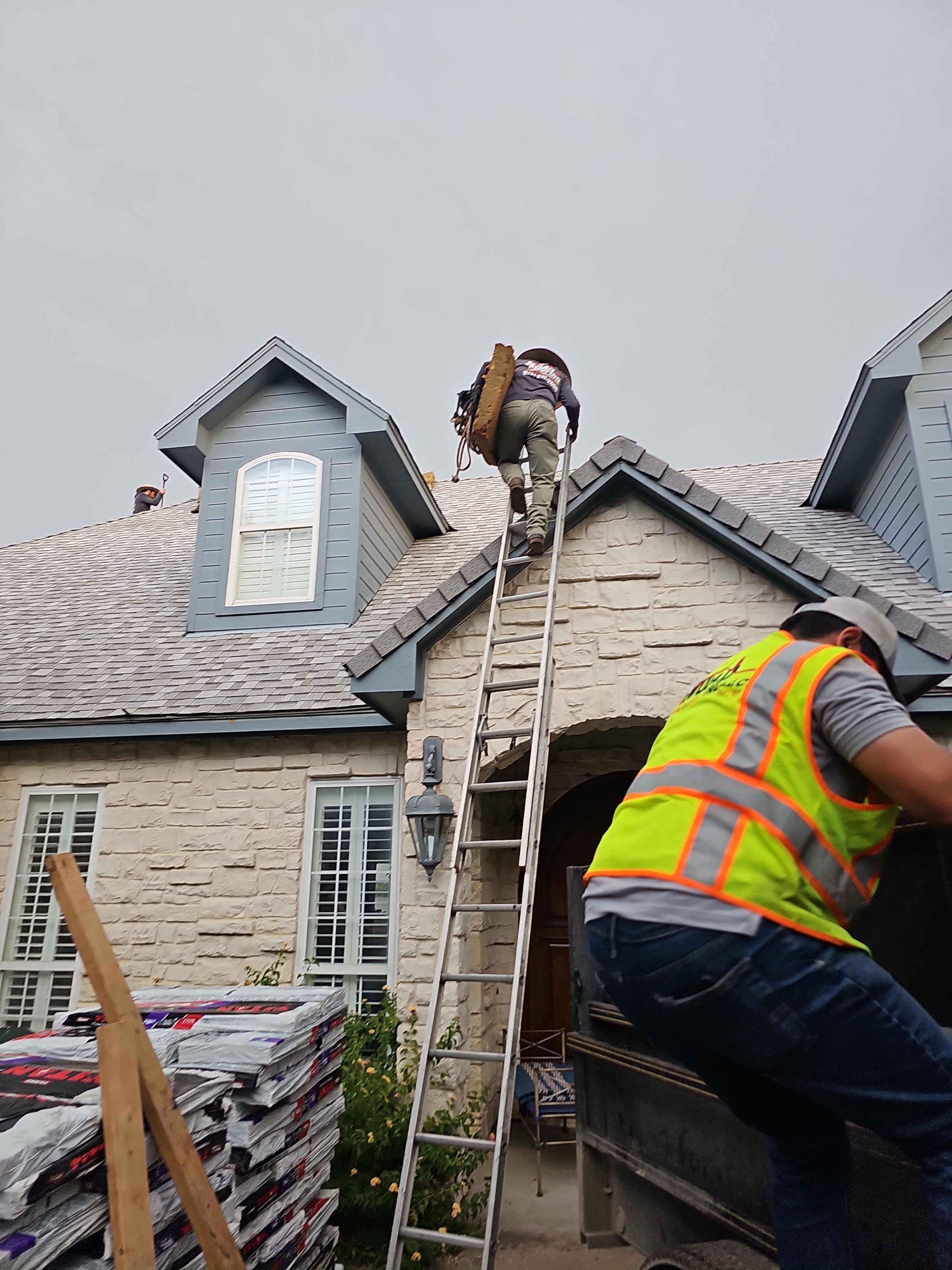 A man in a gray jacket and pants with a large tool on his back, standing on a ladder on the roof of a house, working on the roof. In the foreground, someone is wearing a yellow safety vest, and there are shingles and roofing materials on the ground.