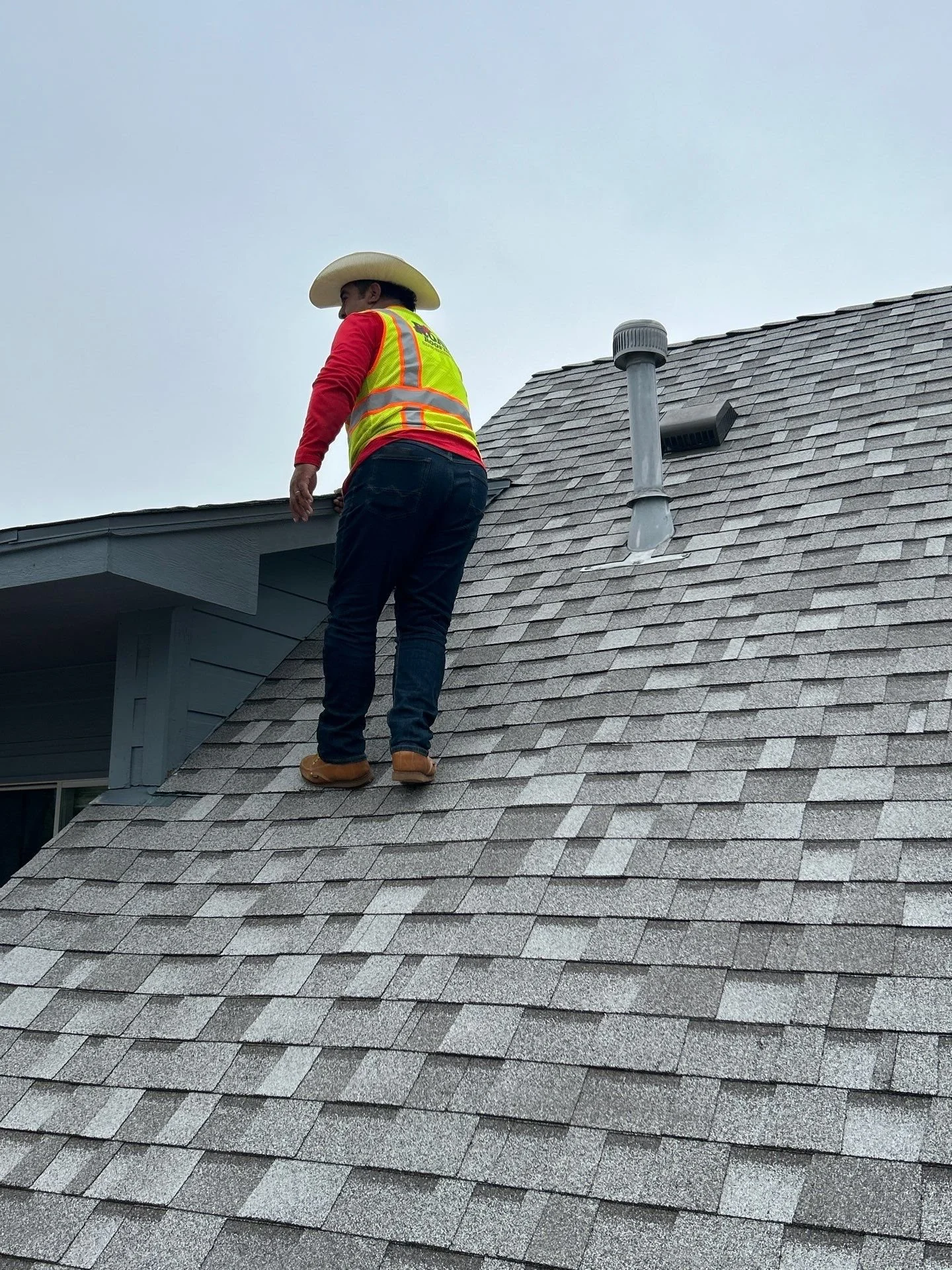 A man wearing a cowboy hat, a high-visibility vest, a red long-sleeve shirt, and jeans stands on the sloped roof of a house, looking downward, with a cloudy sky in the background.