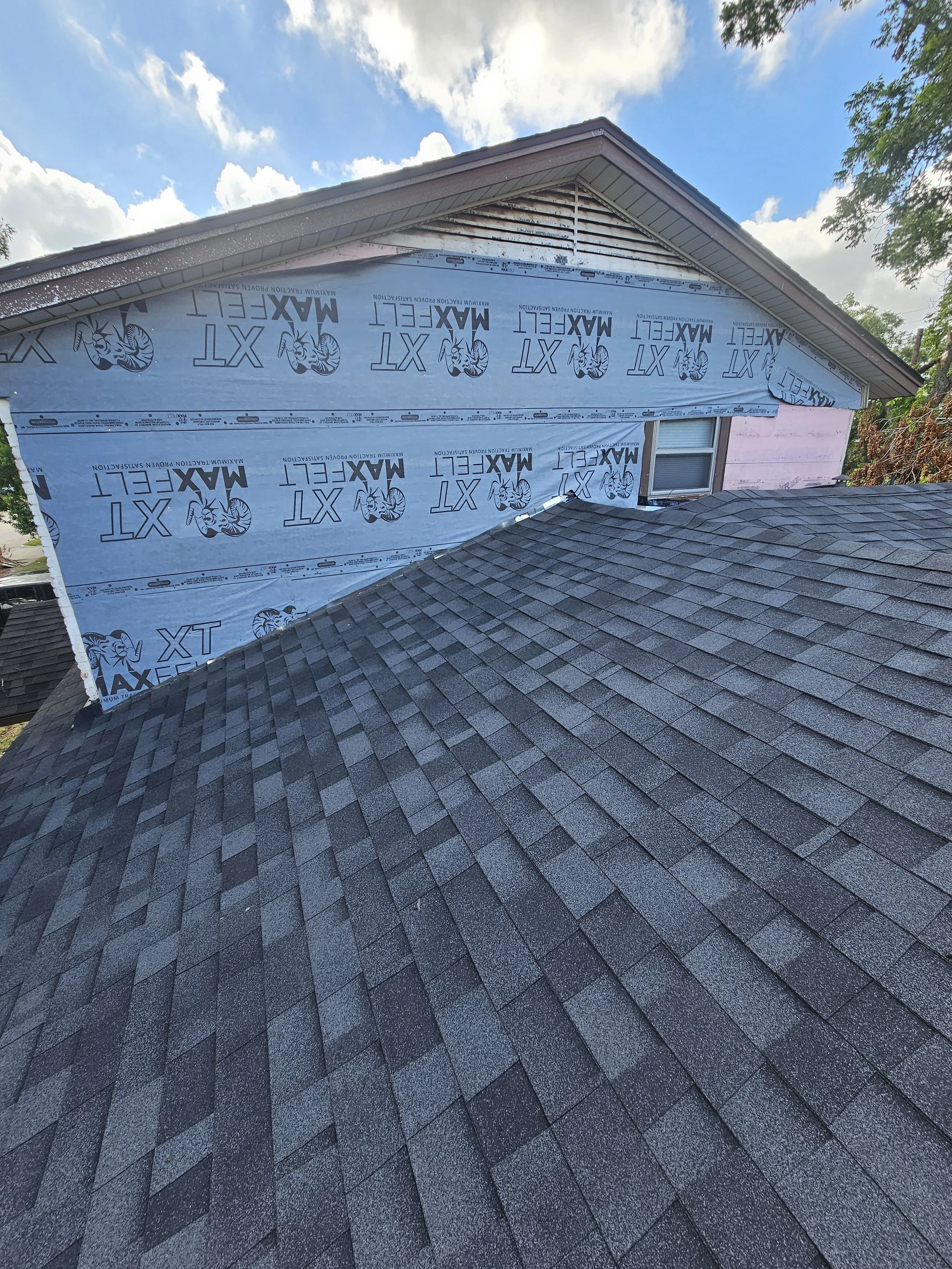 A house under renovation with blue weather-resistant sheathing on the upper part of the exterior wall, and new gray asphalt shingles on the roof. The sheathing has the brand name 'Max-Fel' printed on it with a lobster logo.