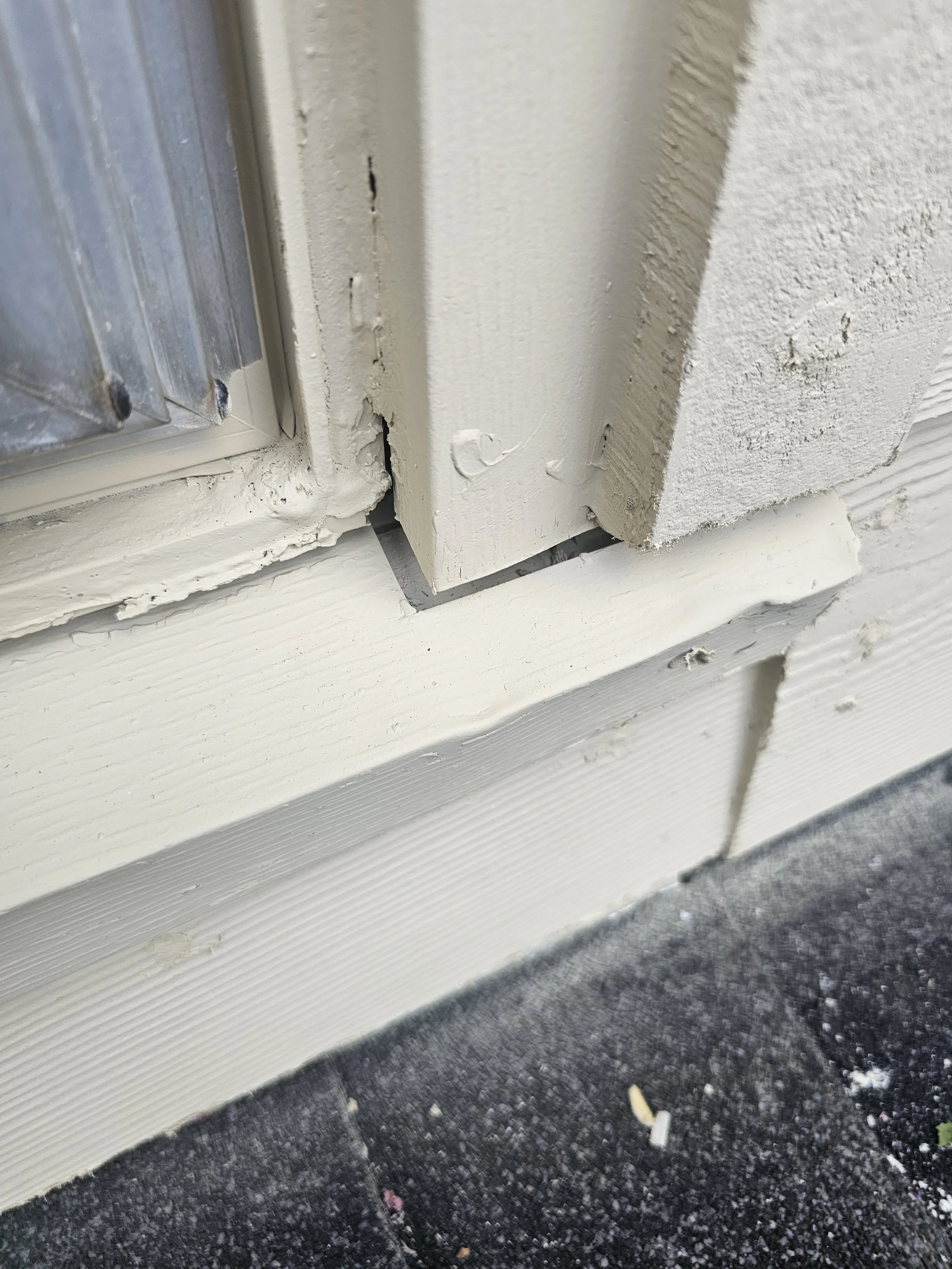 Close-up of a white painted window frame and sill with chipped paint, revealing the wood underneath, resting on a textured dark gray ground with scattered debris.