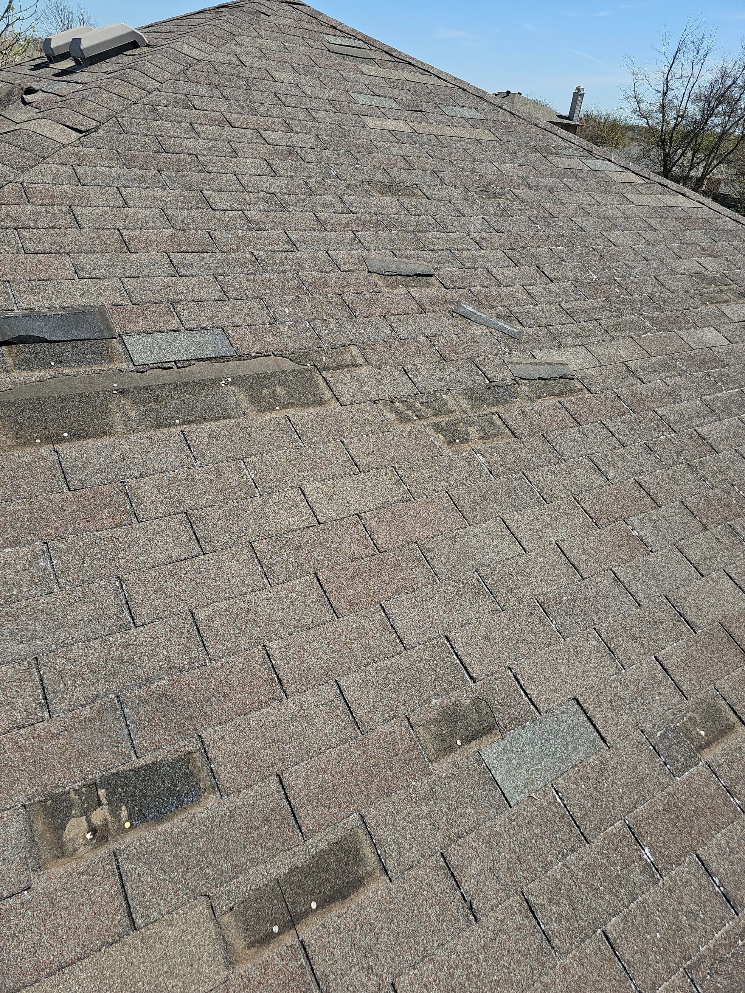 A roof with asphalt shingles, some damaged and missing shingles, under a blue sky with trees in the background.