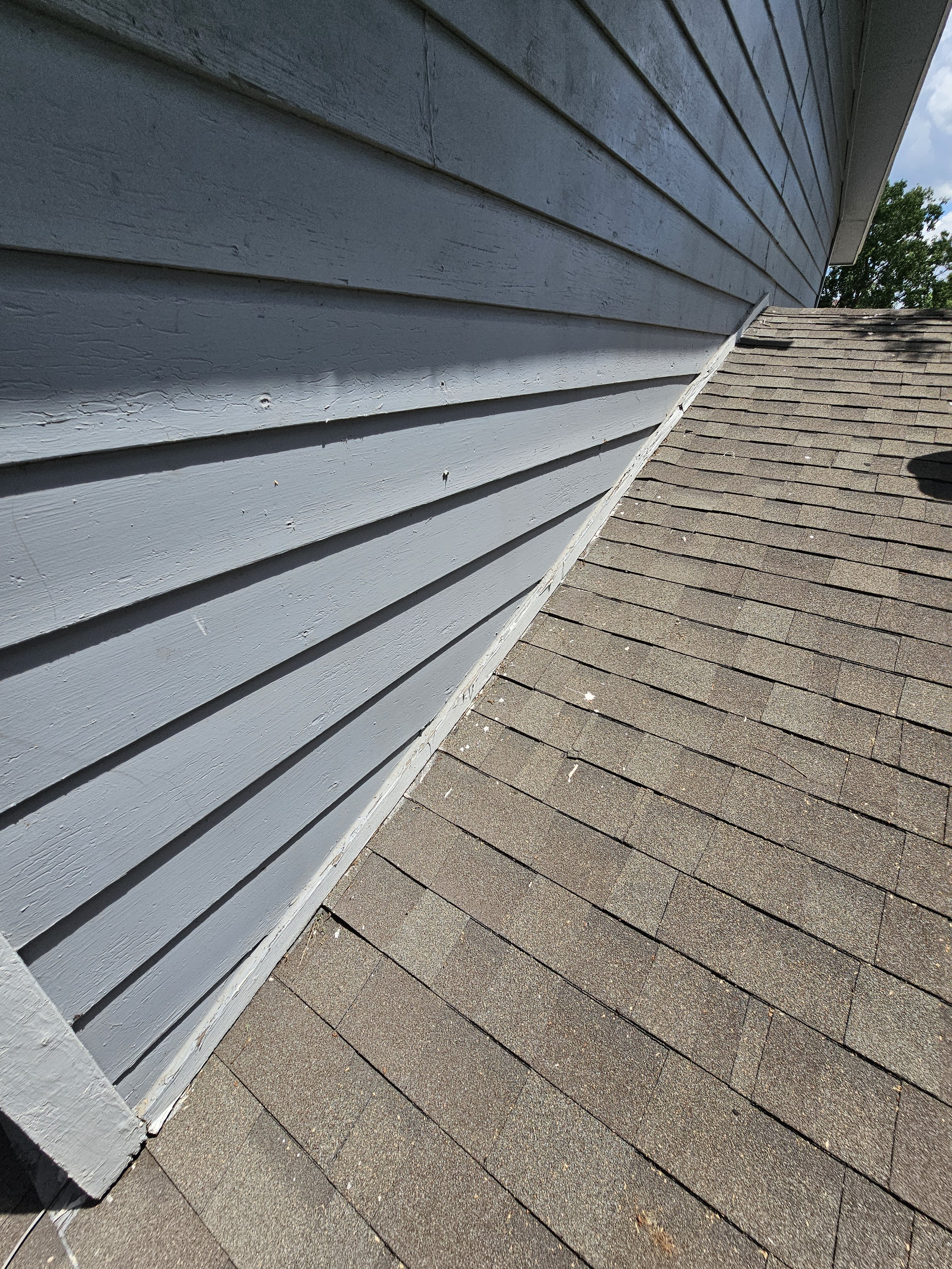 Close-up of the side of a house with gray horizontal wooden siding and a roof with brown asphalt shingles.