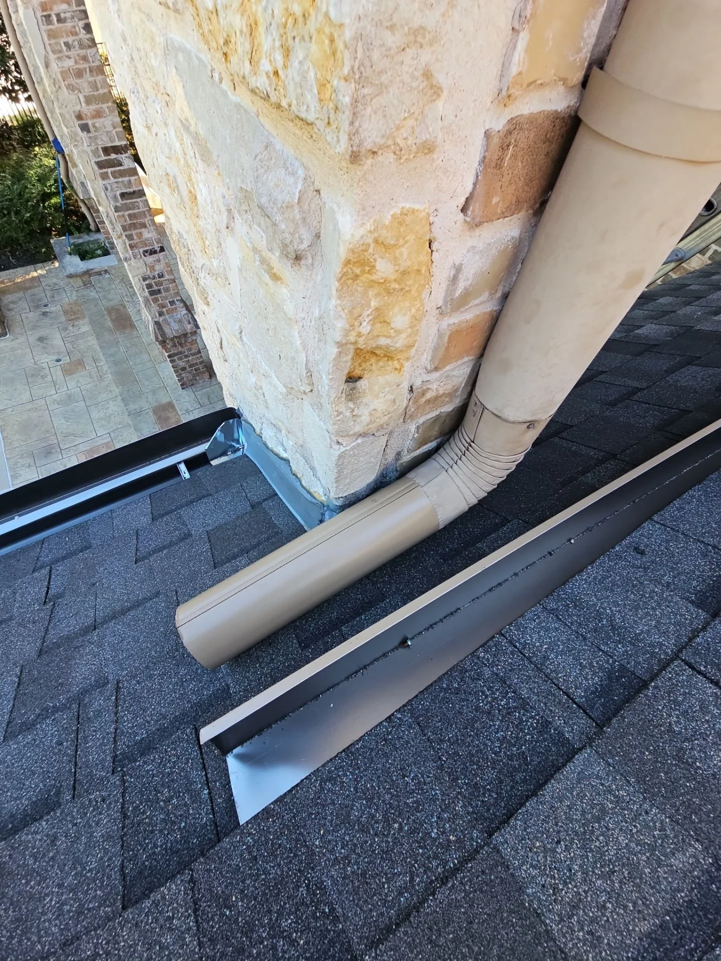 Close-up view of a rooftop with black shingles, a beige downspout drainpipe, and a brick chimney in the background.