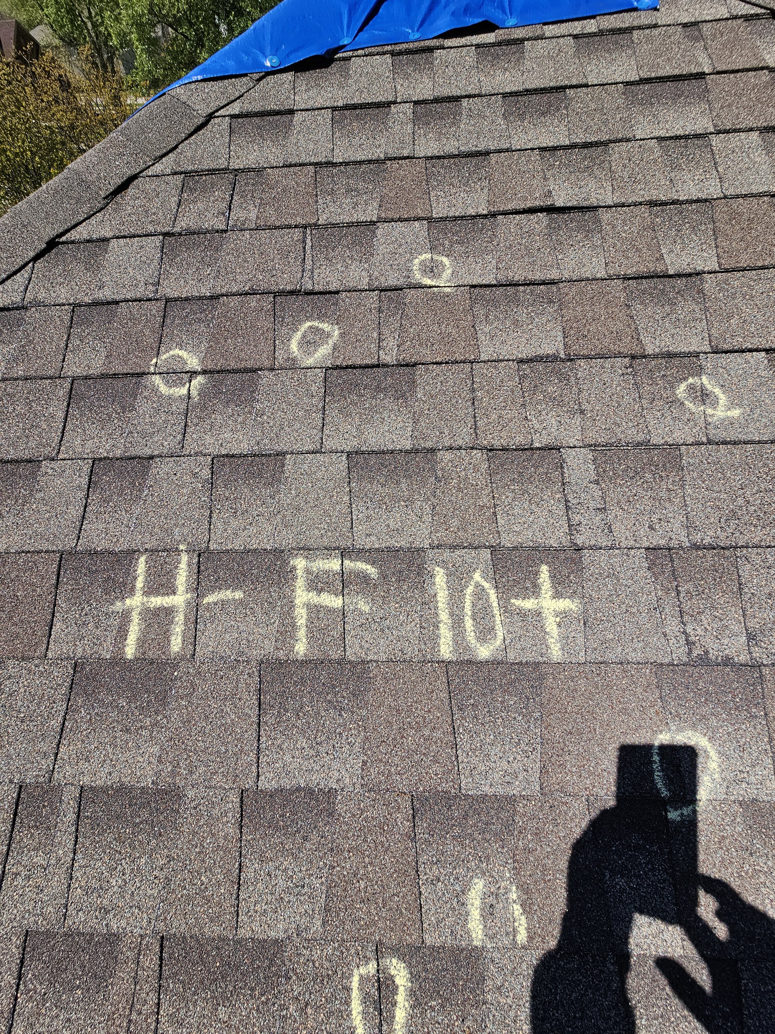 Sunshine illuminating a shingled roof with chalk markings and letters on the surface. A person's shadow and a camera's shadow are visible in the lower right corner.