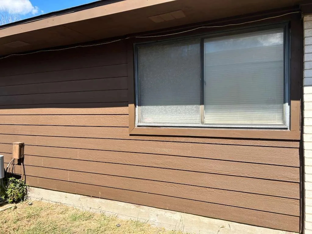 Exterior of a house with brown horizontal siding, a large sliding window with blinds, and a small patch of grass in the yard.