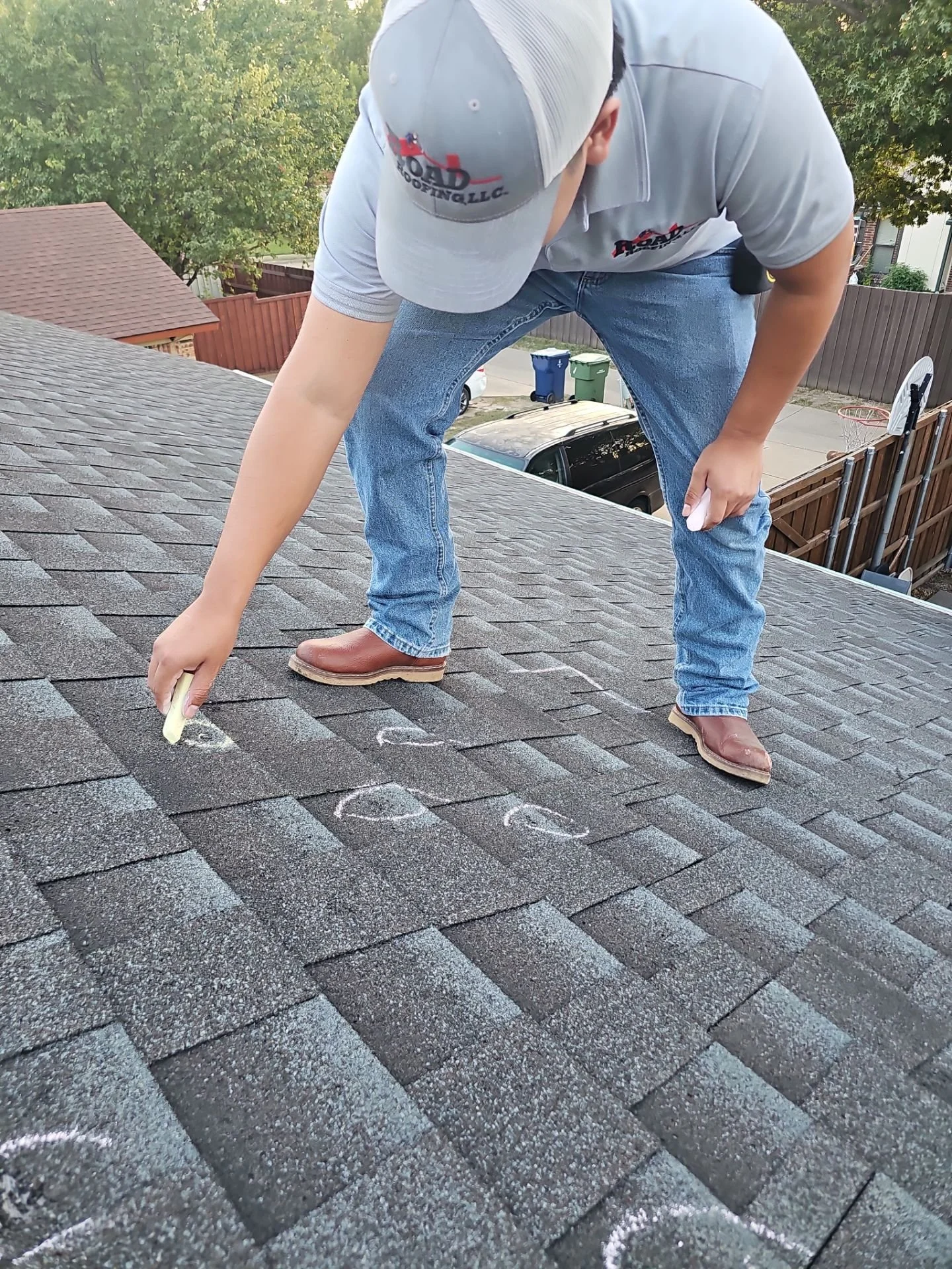 A man in jeans, a gray T-shirt, and a gray cap is kneeling on a roof, using chalk to draw on the shingles.