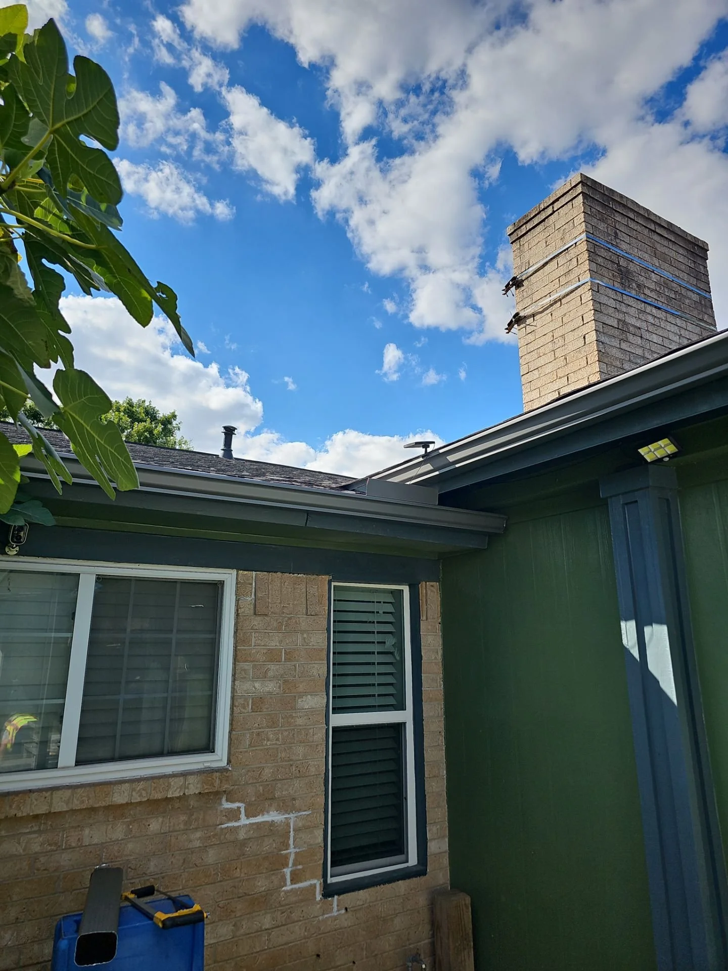 The image shows the side of a house with a brick chimney, a window with white blinds, and a brick wall part. There's a dark green painted porch or wall area and a shop vacuum or similar tool on a blue container. The sky above is partly cloudy with bl