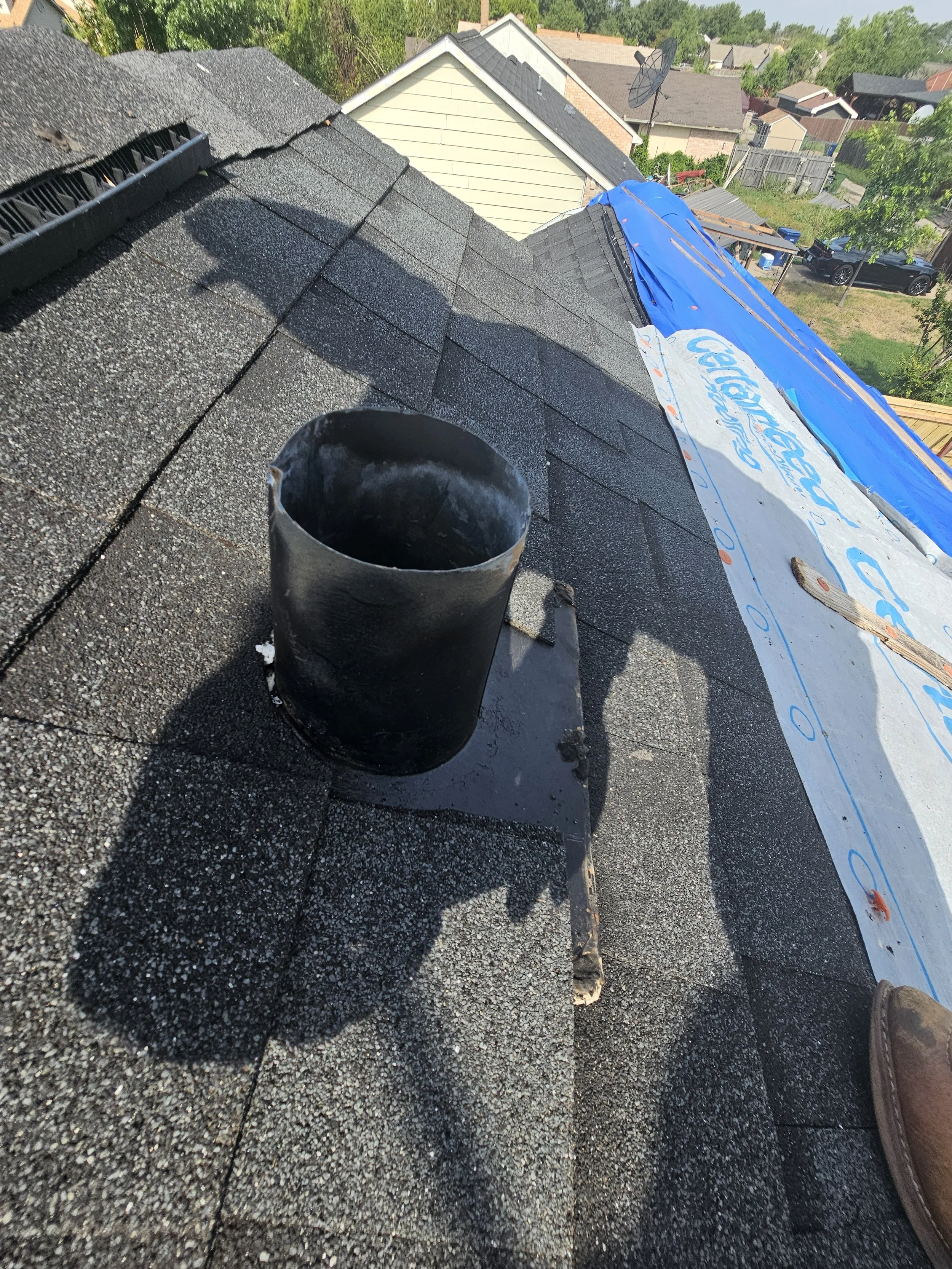View of a roof under construction with new asphalt shingles, a black vent pipe, and part of a house with a white fence, trees, and neighboring houses in the background.