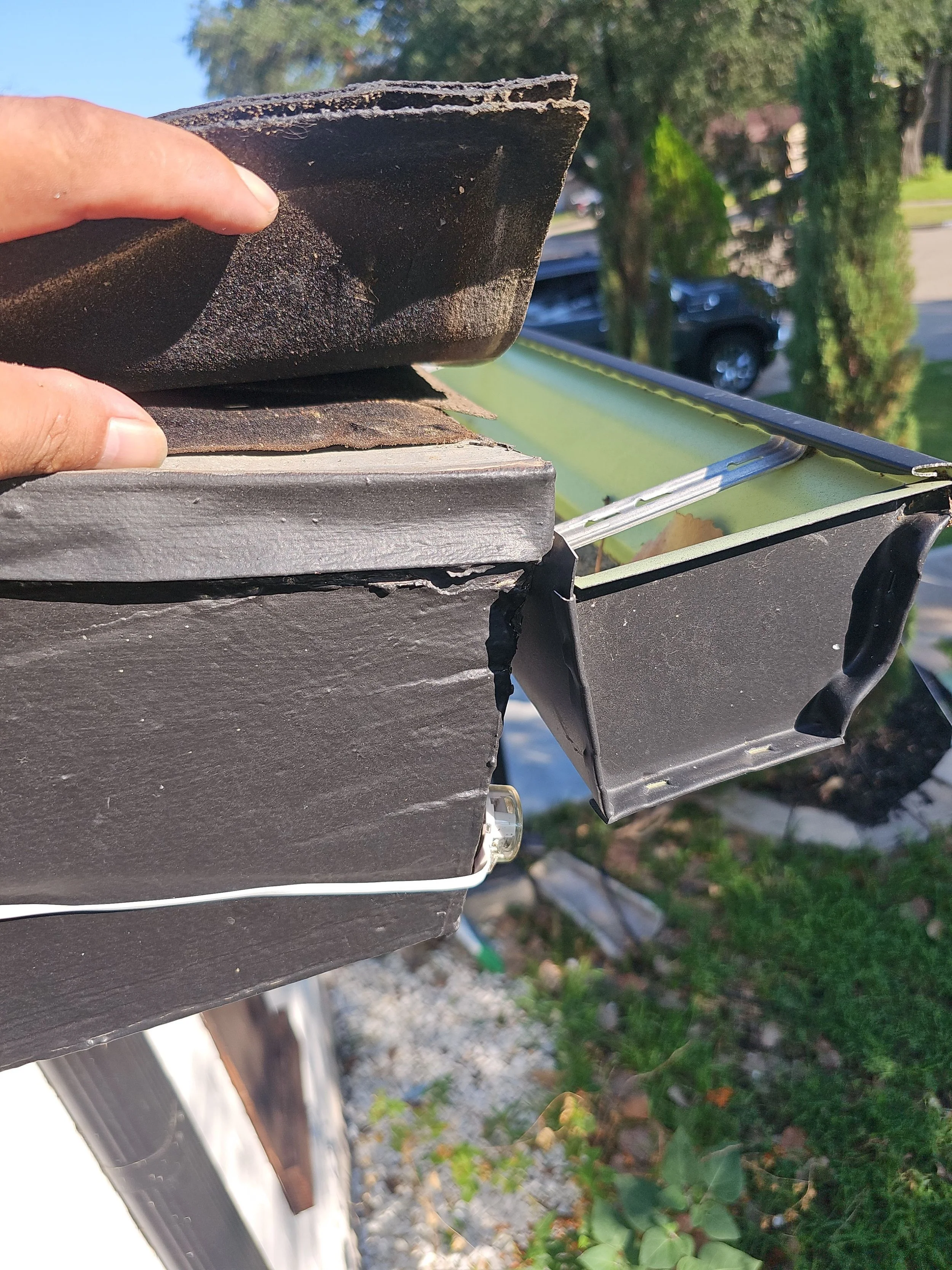 Close-up of a person installing or repairing roof flashing on a house gutter, with a background of trees, grass, and a parked vehicle.