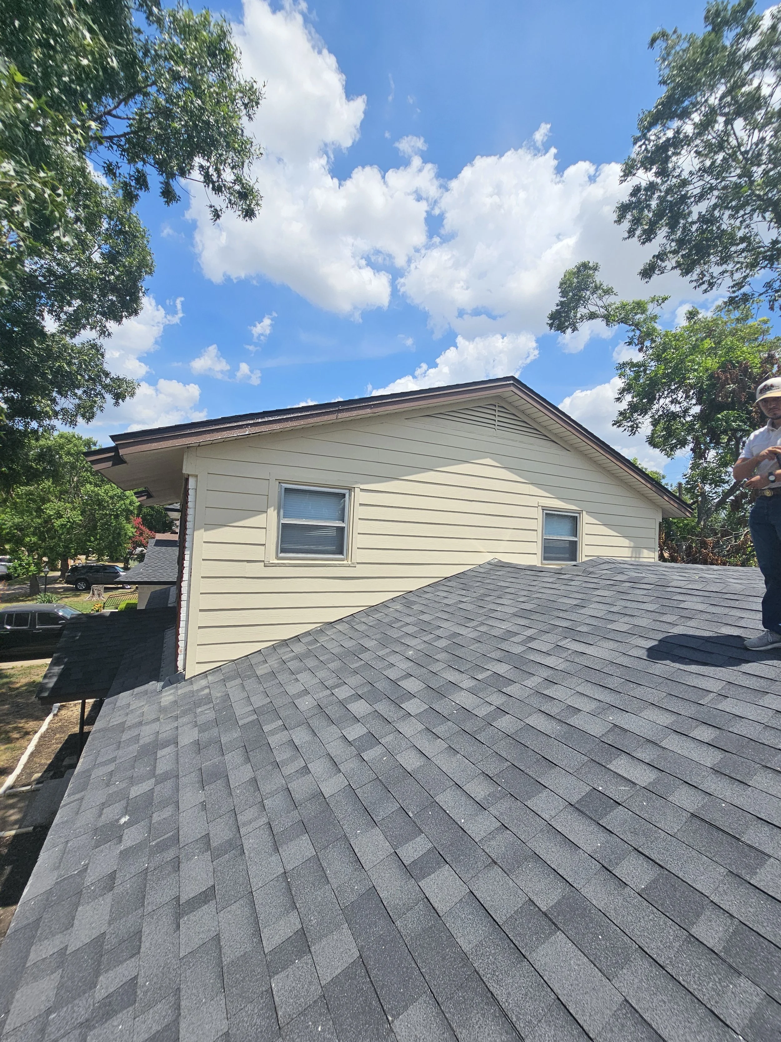 View of the roof of a house with dark asphalt shingles, side of the house with beige siding, and a man standing near the edge of the roof, with trees and a blue sky with fluffy clouds in the background.