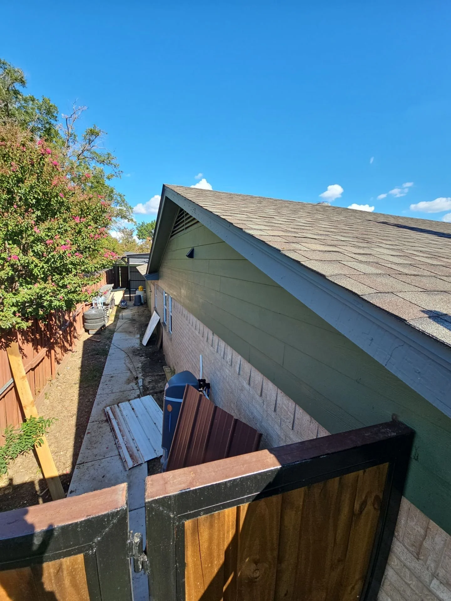 View of a backyard with a sloped roof of a house, trees with pink flowers, a wooden fence, and various construction materials and tools along a narrow walkway.