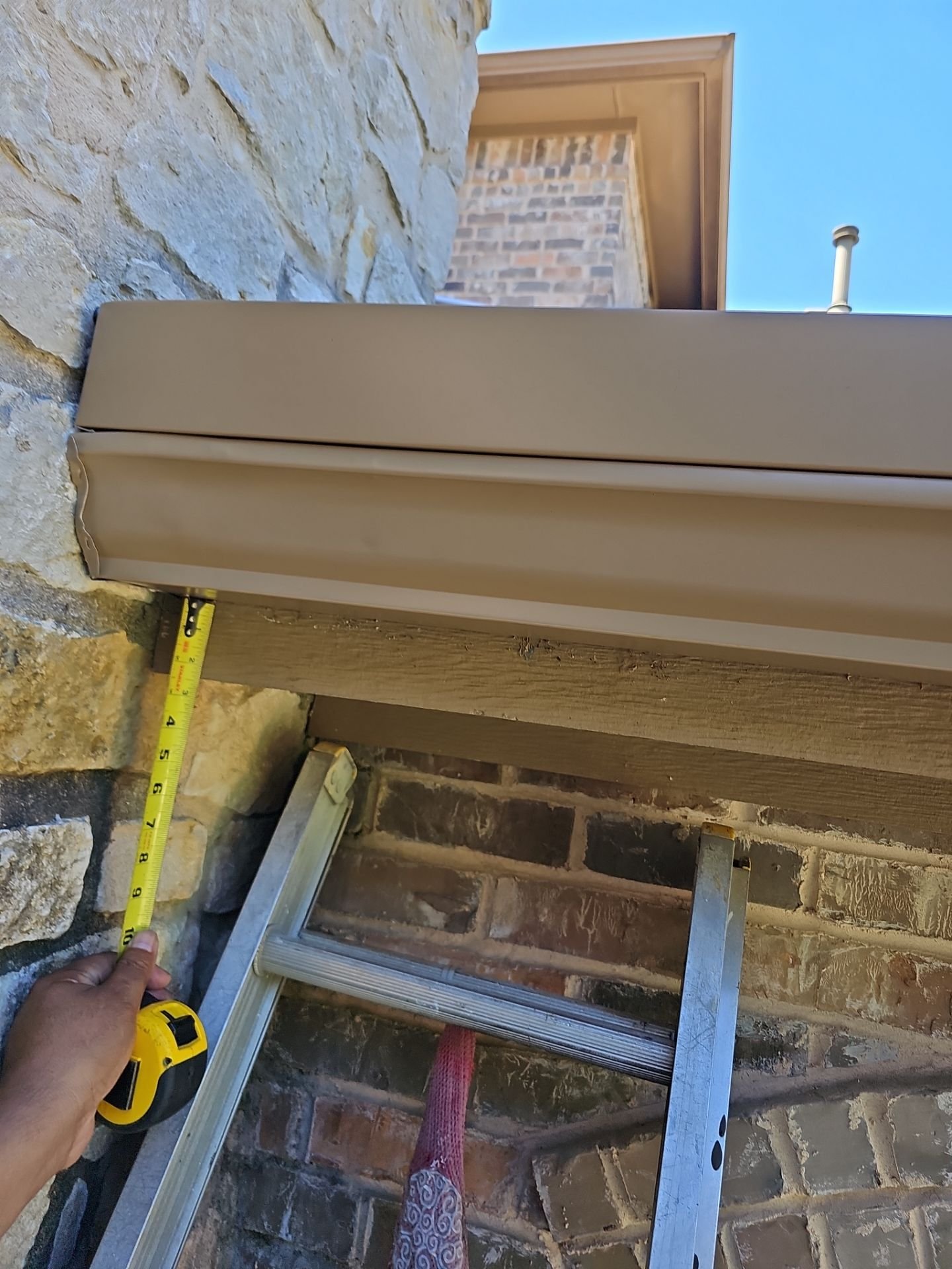 A person holding a measuring tape next to a ladder on a decorated brick and stone outdoor wall. Part of a beige box or panel is visible, along with a window and a roof overhang in the background.