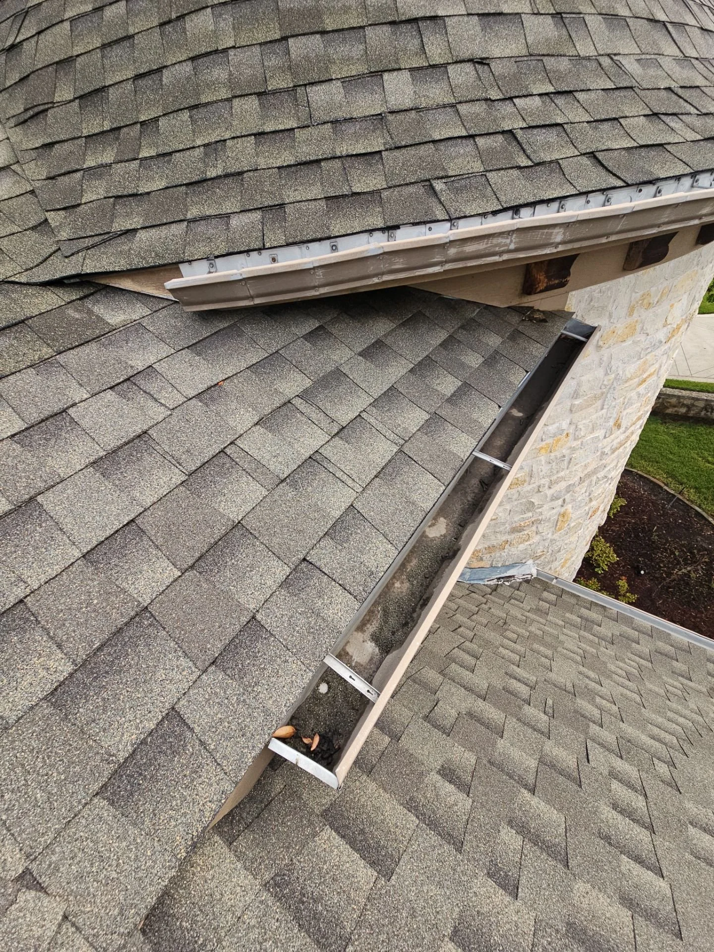 Close-up of a roof with asphalt shingles, a gutter, and a gutter guard. The roof has brown and gray shingles, some of which are slightly curled or damaged. The gutter runs along the edge of the roof and contains some dirt and debris.