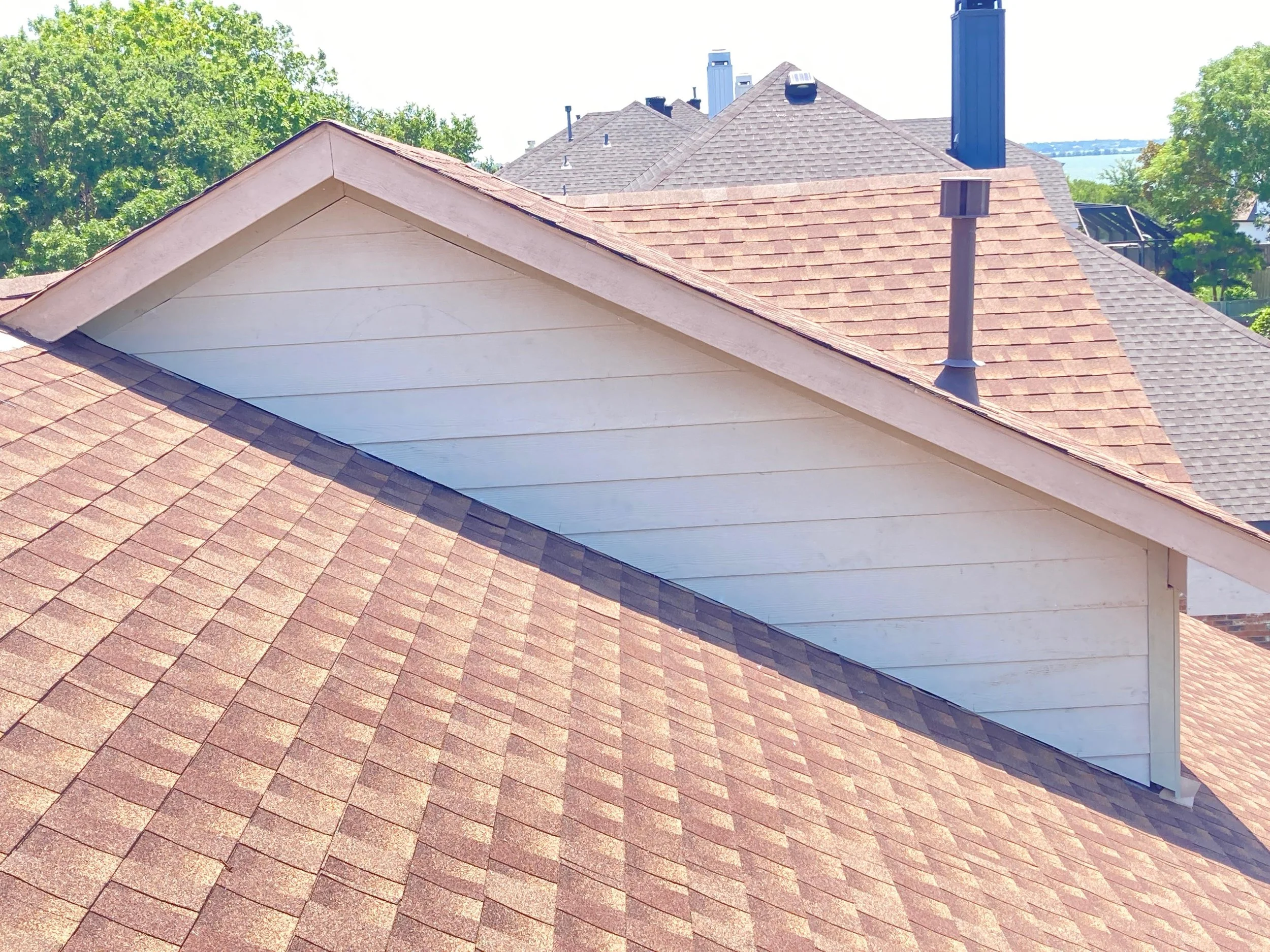 View of multiple house rooftops with different shingles, some are reddish-brown, and others are gray, during daytime with green trees in the background and clear blue sky.