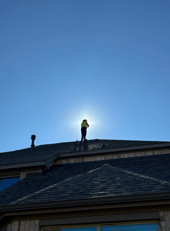 Person standing on the roof of a house during daytime with the sun behind them, creating a halo effect.