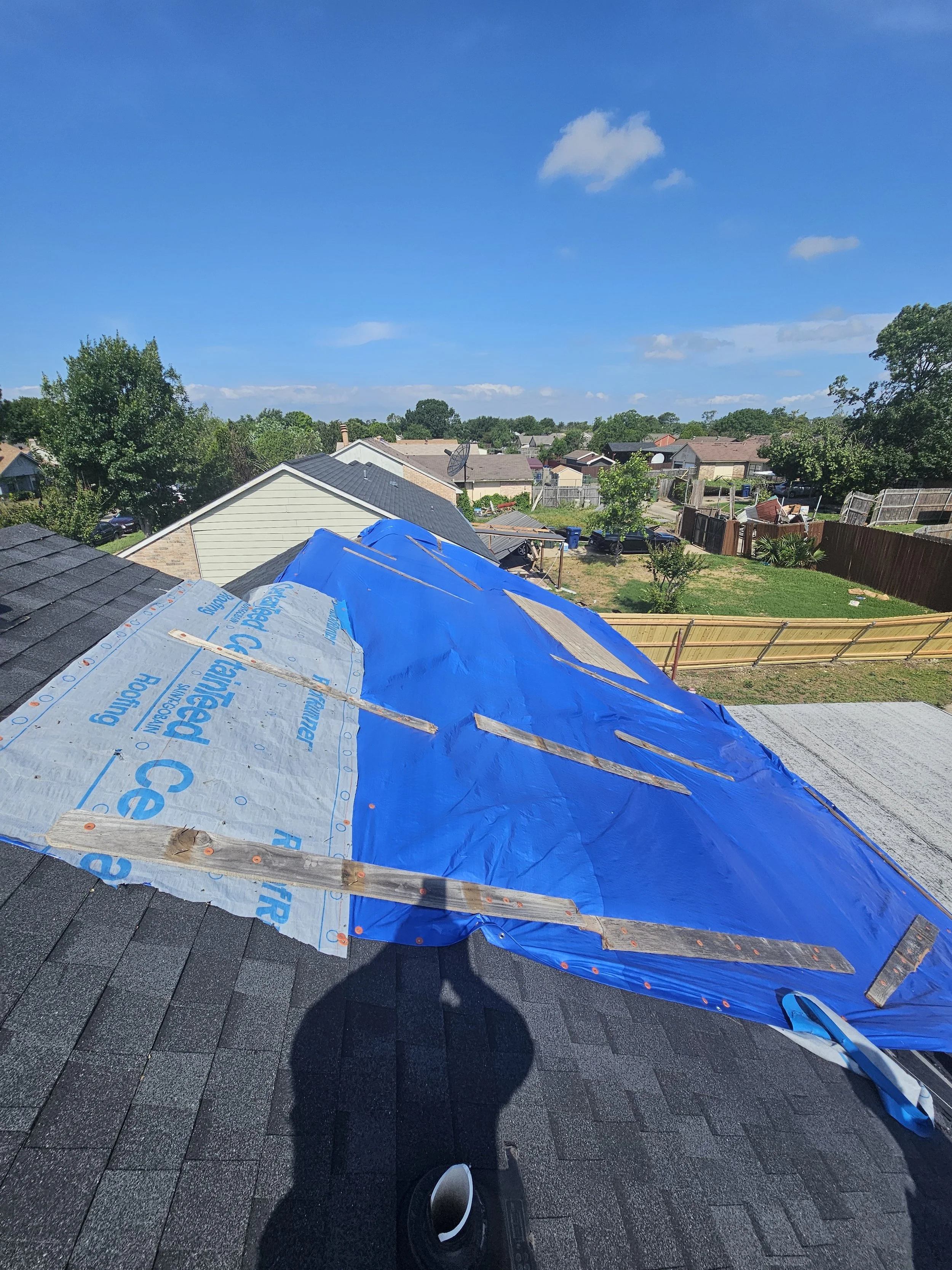 View of a residential backyard with a house's roof under construction, unfinished roofing materials and blue tarps, neighboring houses, a clear blue sky with scattered clouds, and a person's shadow and footwear visible in the foreground.