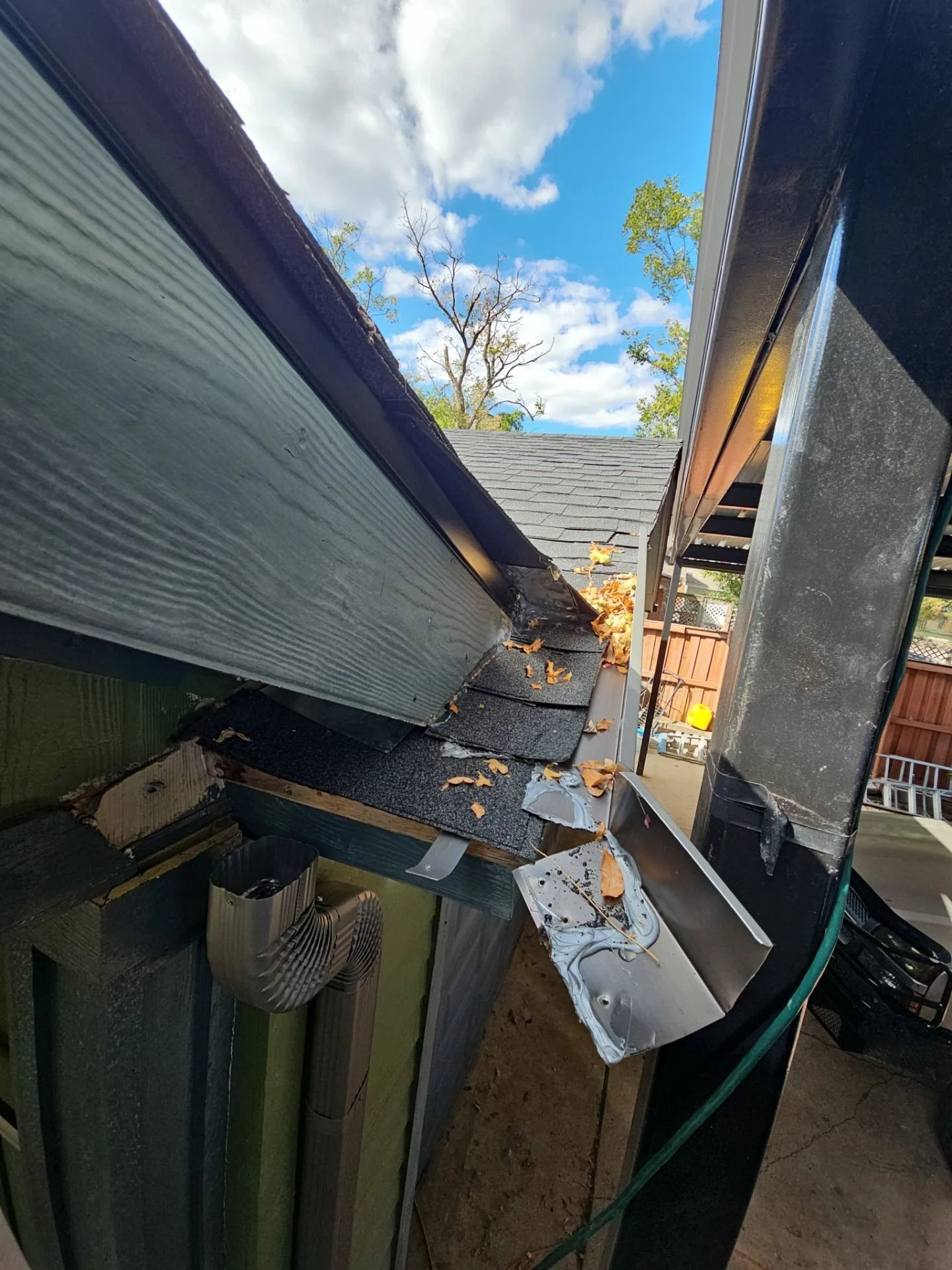 View of a damaged house gutter with leaves and debris inside, under a blue sky with clouds and some trees visible in the background.