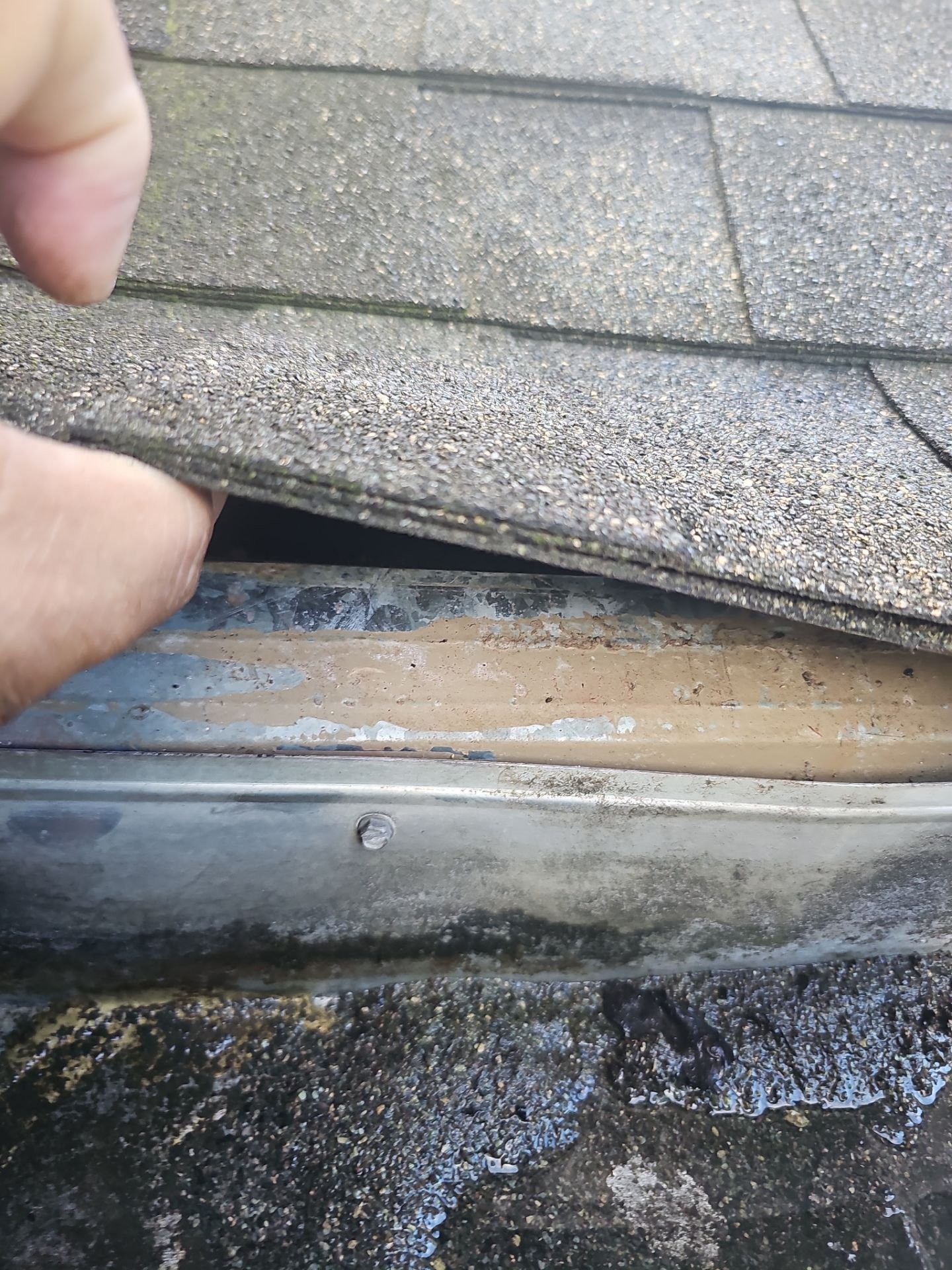 Close-up of a person's fingers lifting a loose shingle on a roof, revealing underlayment and the edge of the roof's surface, with shingles and construction debris visible below.