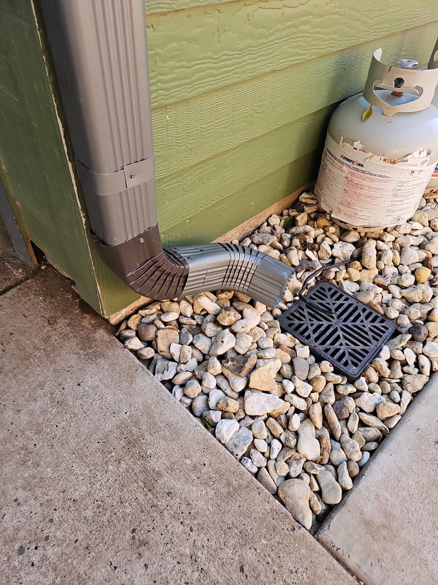 An outdoor scene showing a green wooden wall with a metal drainpipe and extension, a propane tank, a black drain cover, a concrete sidewalk, and a gravel area with small stones.