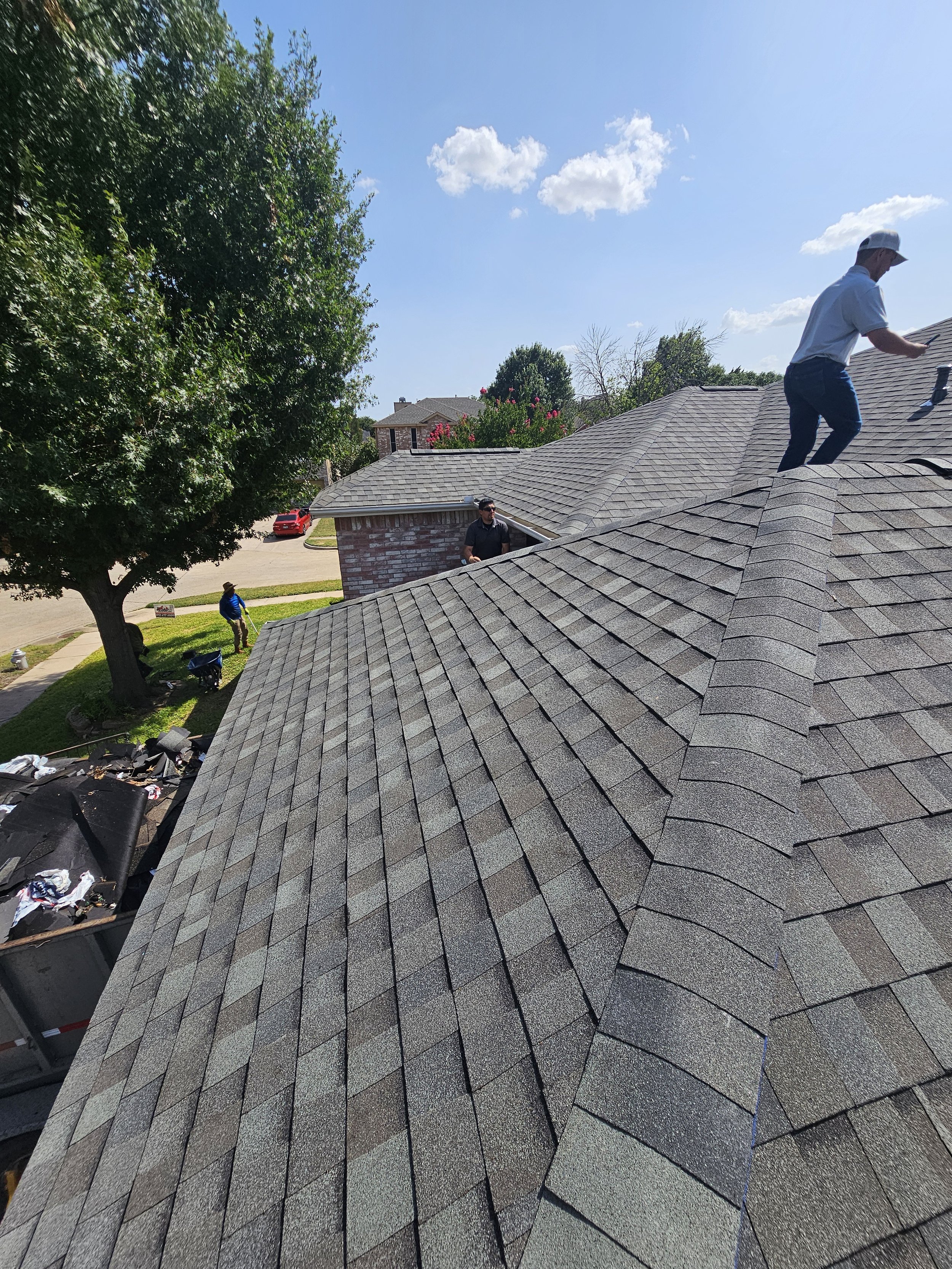 Two workers on a residential roof installing new asphalt shingles under a clear blue sky with some clouds. One worker is near the chimney and another near the peak of the roof, and there are trees and houses visible in the background.