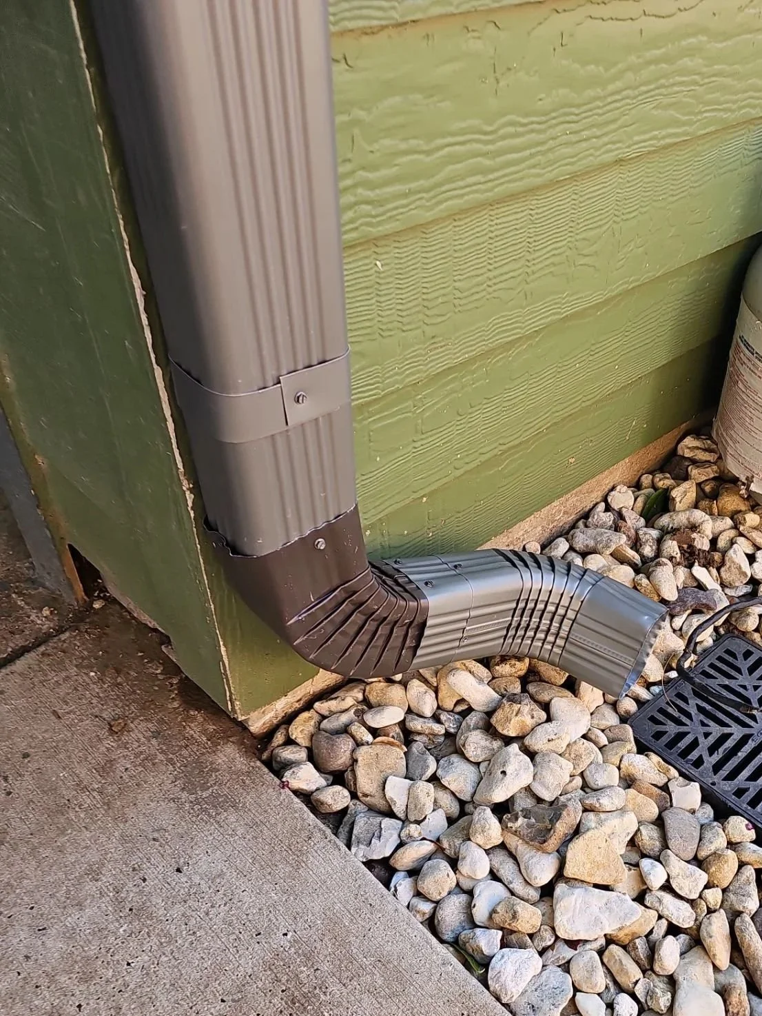 A gray metal downspout connected to a rain gutter runs along the corner of a green wooden building, ending above a bed of light-colored rocks near a black drainage grate.
