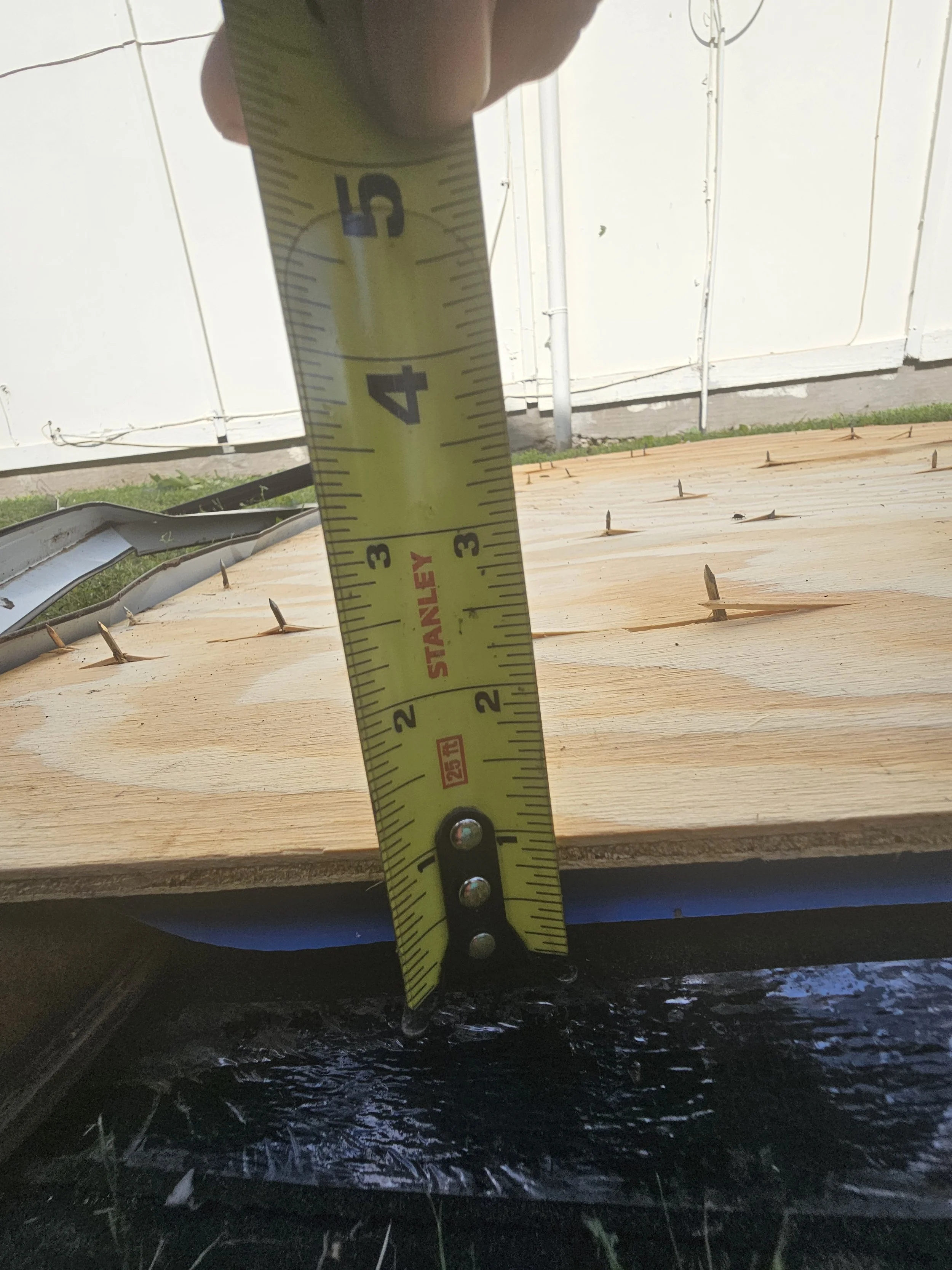 Close-up of a yellow measuring tape showing approximately 4 inches in height, positioned on a wooden surface with nails, in front of a white wall outdoors.