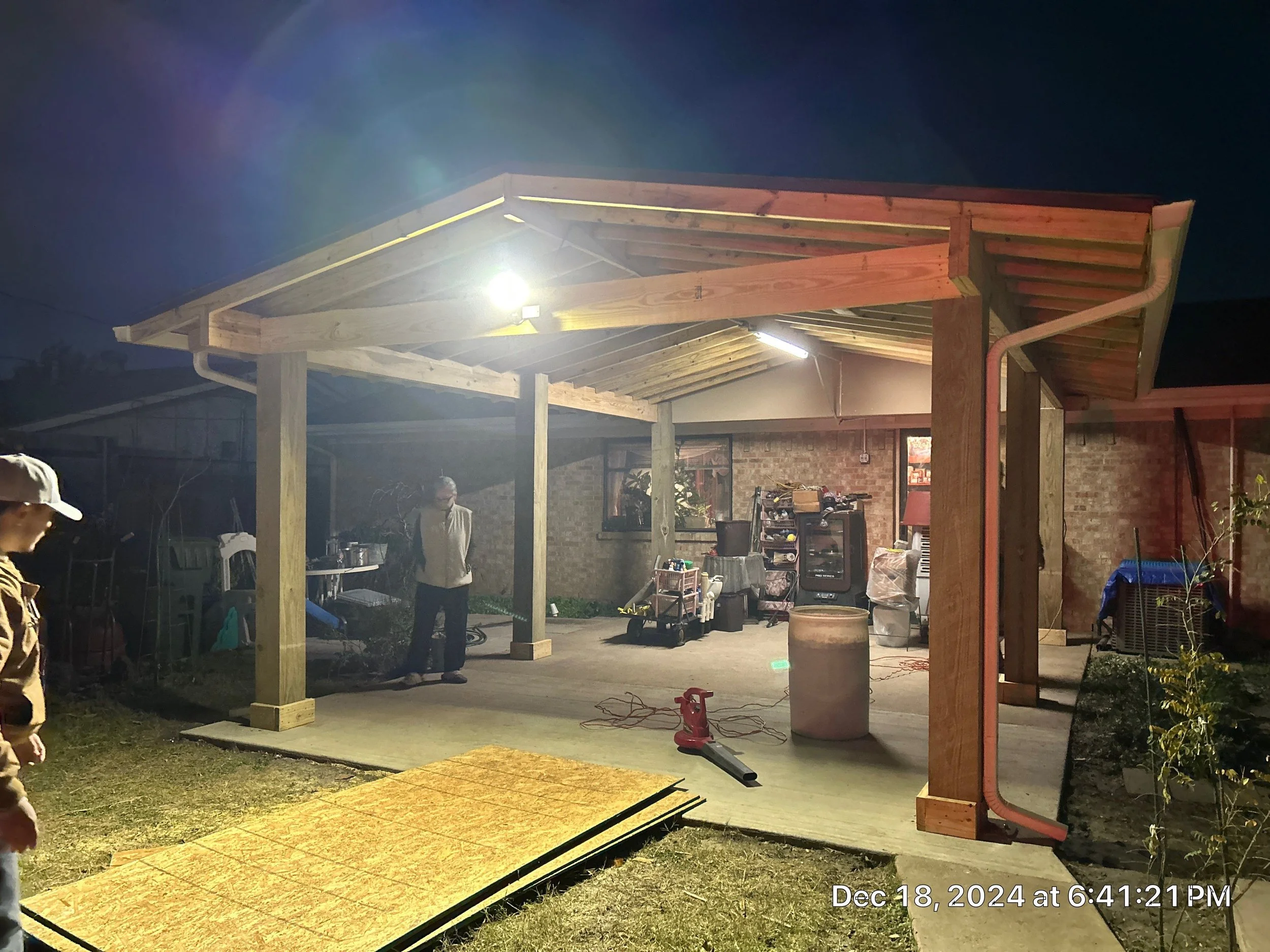 This image shows a backyard construction scene at night. There is a wooden structure with a roof frame being built, illuminated by bright work lights. Several people are present, some standing and others working near the wooden frame. Construction to