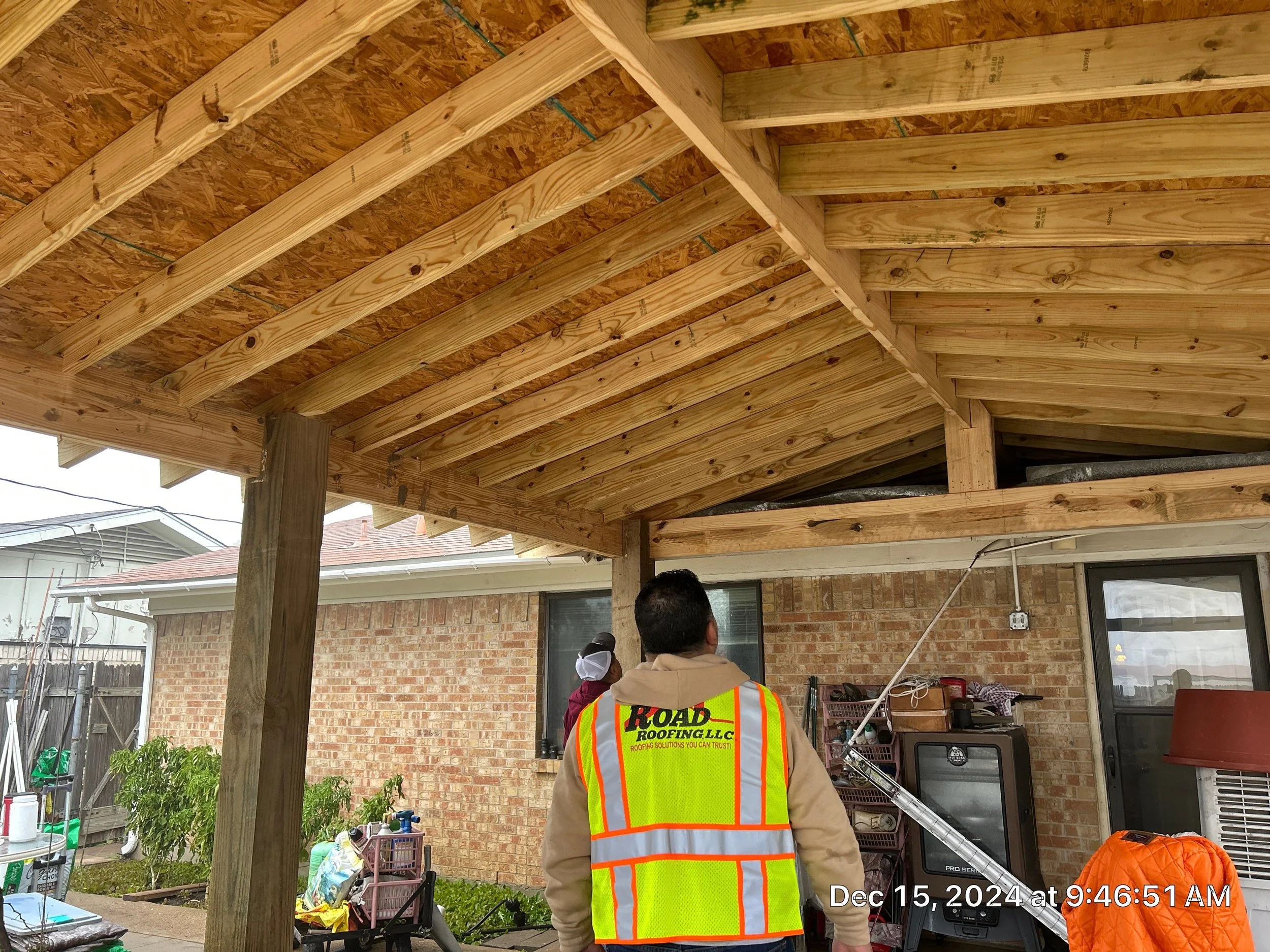 Construction workers installing a wooden roof structure over a patio area, with one worker wearing a bright orange safety vest with the logo 'ROAD ROOFING, LLC.' and a man in a tan jacket and cap. The setting appears to be a backyard with brick walls
