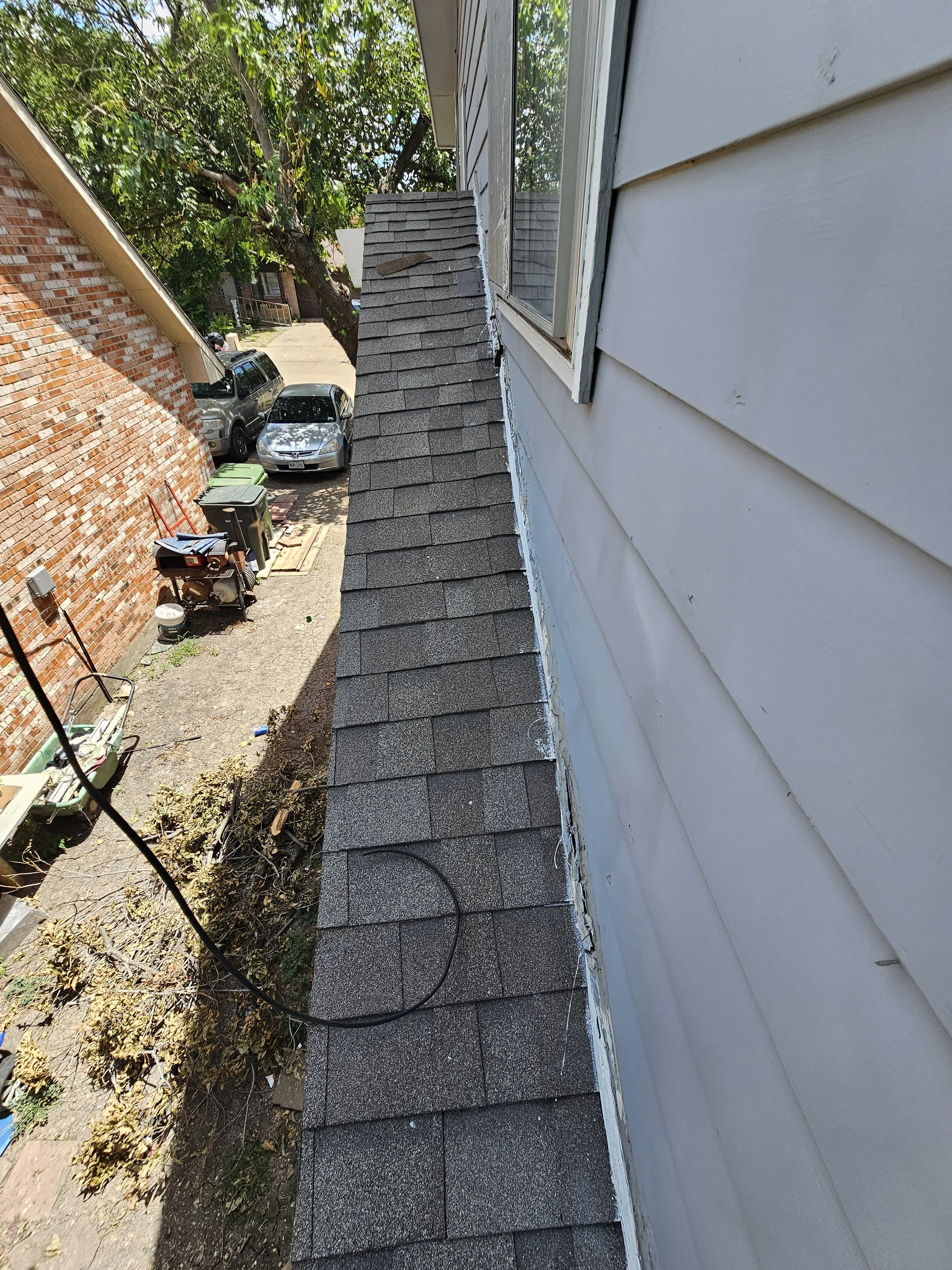 A view from the roof of a house looking down the side wall and neighborhood street, showing parked cars, trash bins, trees, and a neighboring house.