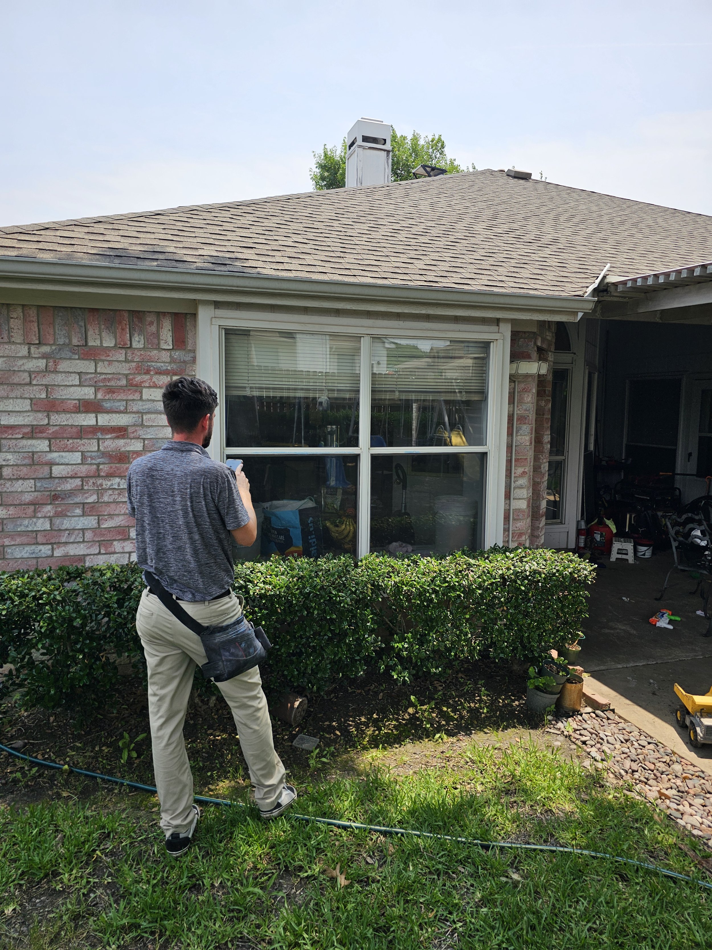 A man in beige pants and a gray shirt is standing outside near a green hedge, looking at his phone, with a house in the background. The house has a brick wall, large window with blinds, and a sloped shingle roof with a chimney. The yard has some gard