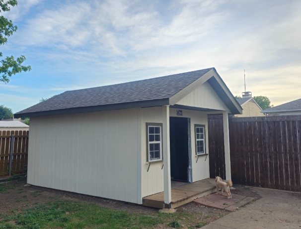 Small beige backyard shed with a dark gray roof, two small windows, an open door, and a small dog standing on the wooden porch in a fenced backyard.