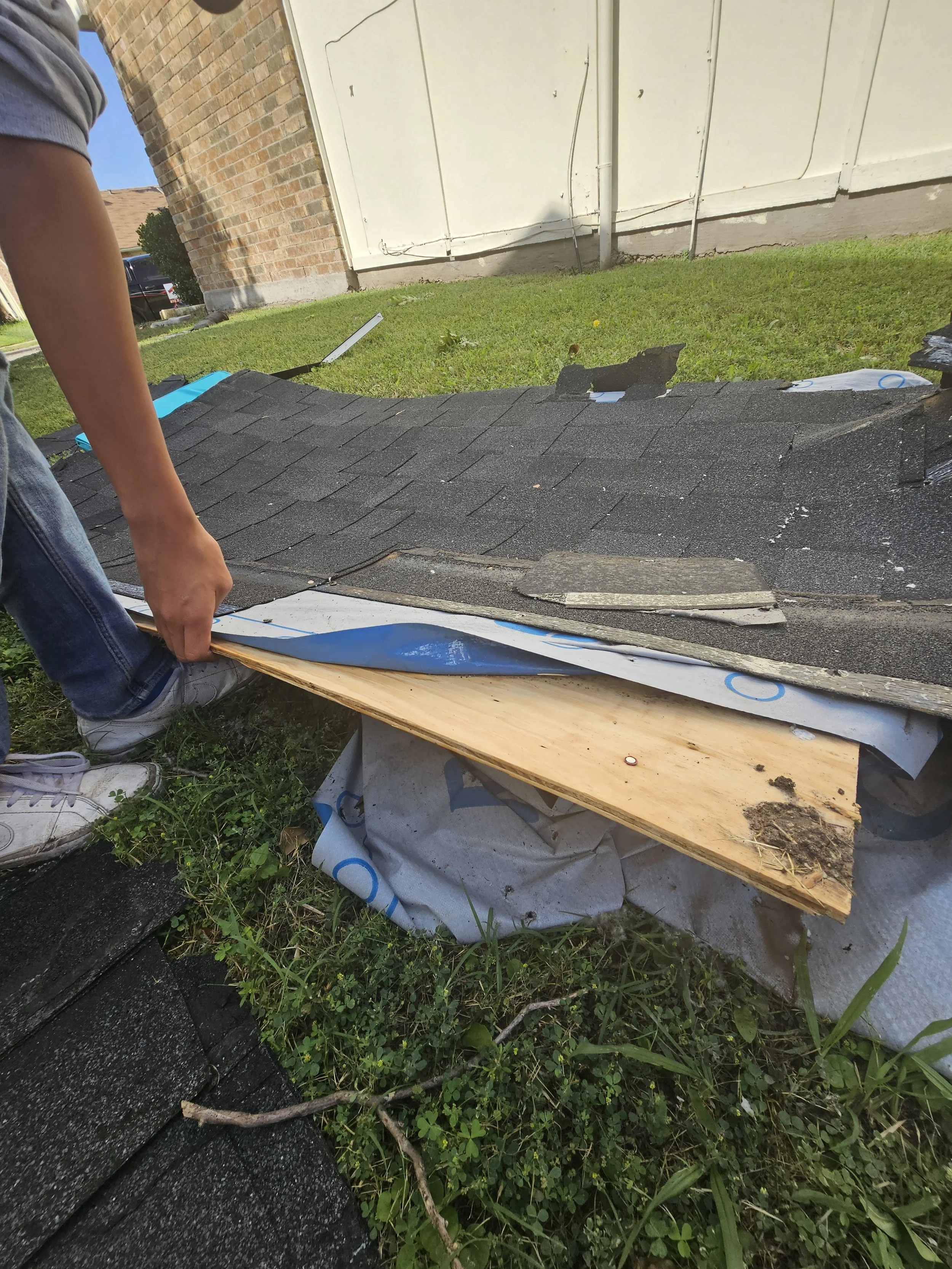 A person installing black shingles on a roof, with green grass and a brick wall in the background.